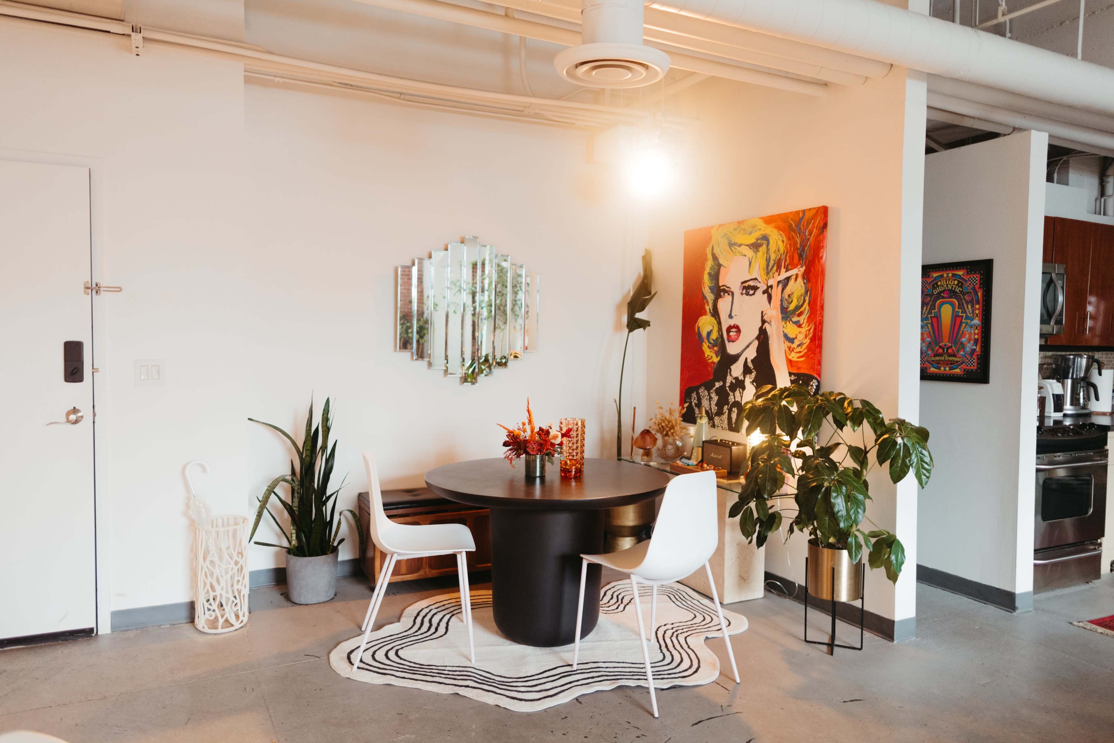 A dining area features a round black table surrounded by two white chairs, with a colorful wall art piece and potted plants nearby.