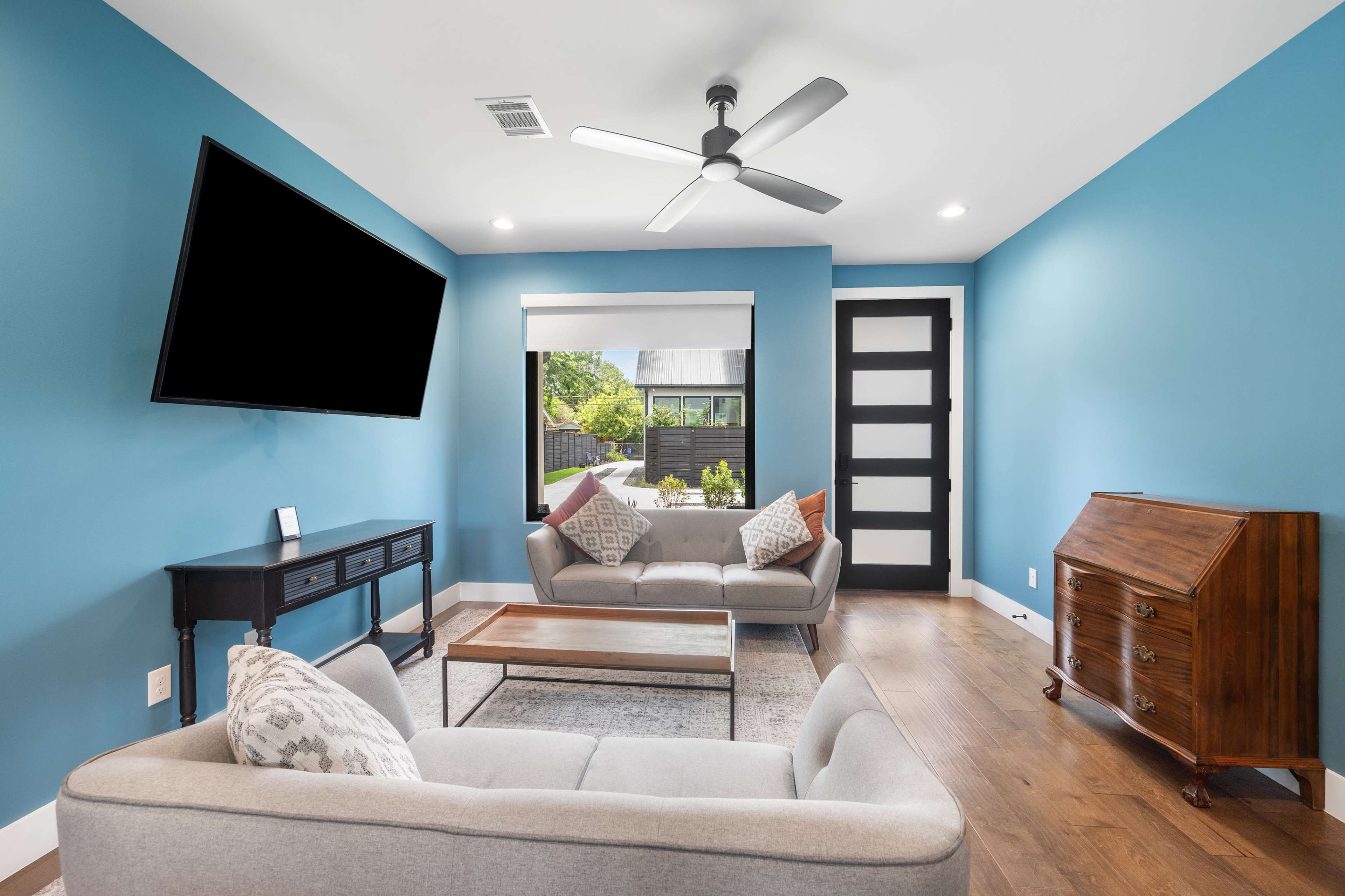 The image shows a modern living room with blue walls, a ceiling fan, a television mounted on the wall, and a seating area featuring a light-colored sofa and a wooden side table.