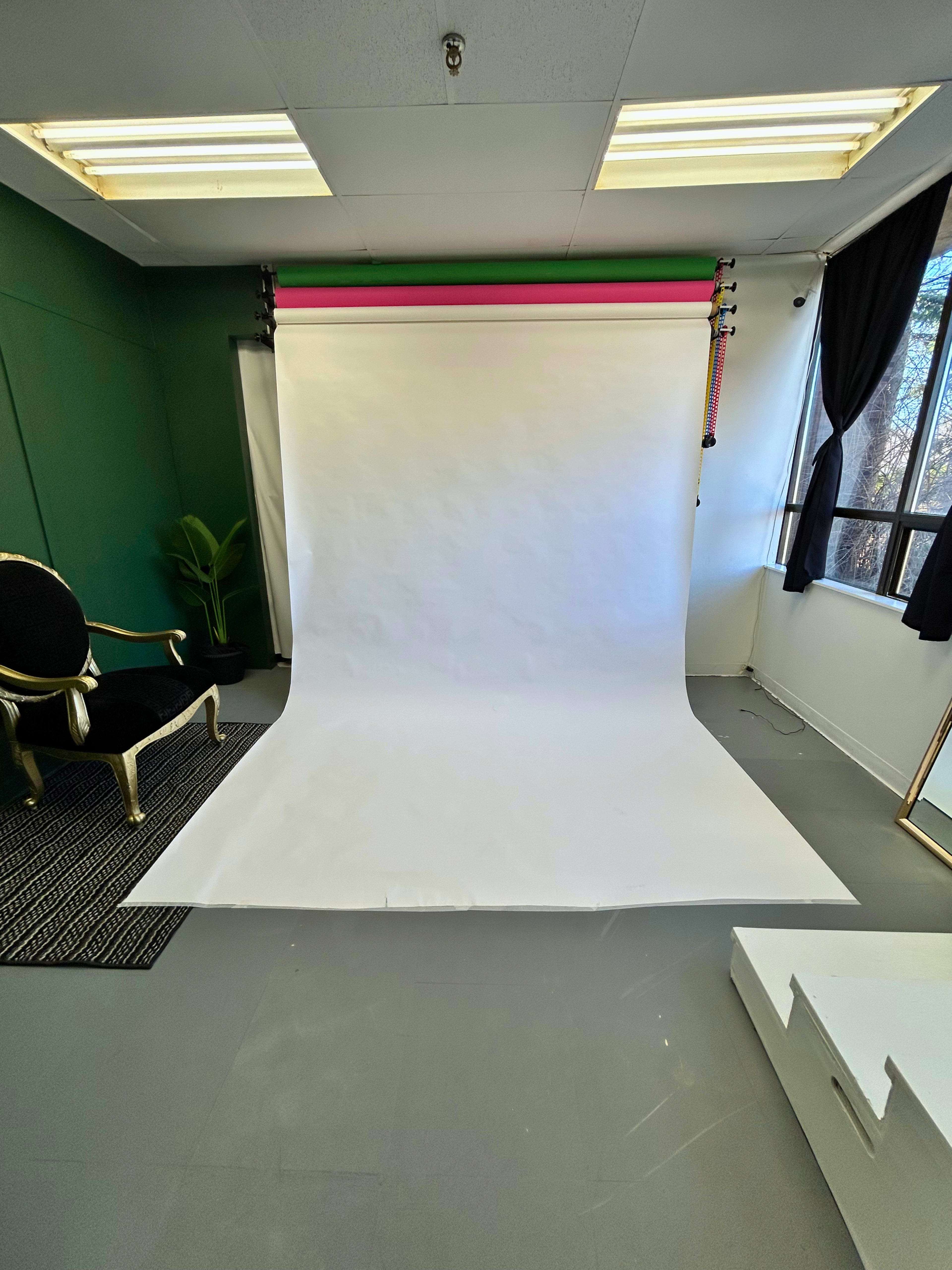 The image shows a photography studio with a white backdrop, green walls, and a black chair positioned on a patterned rug.