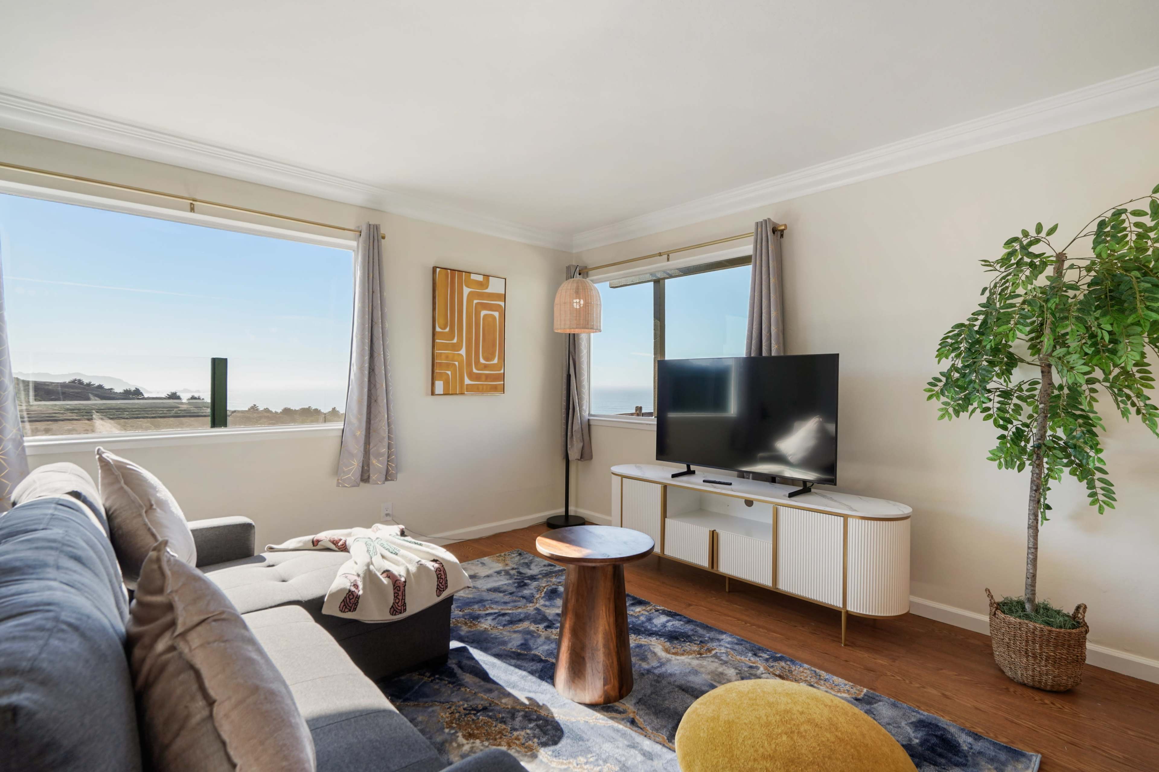 The living room features a gray sofa, a round wooden coffee table, a large television on a white console, and a potted plant near the window with a view of the outdoors.