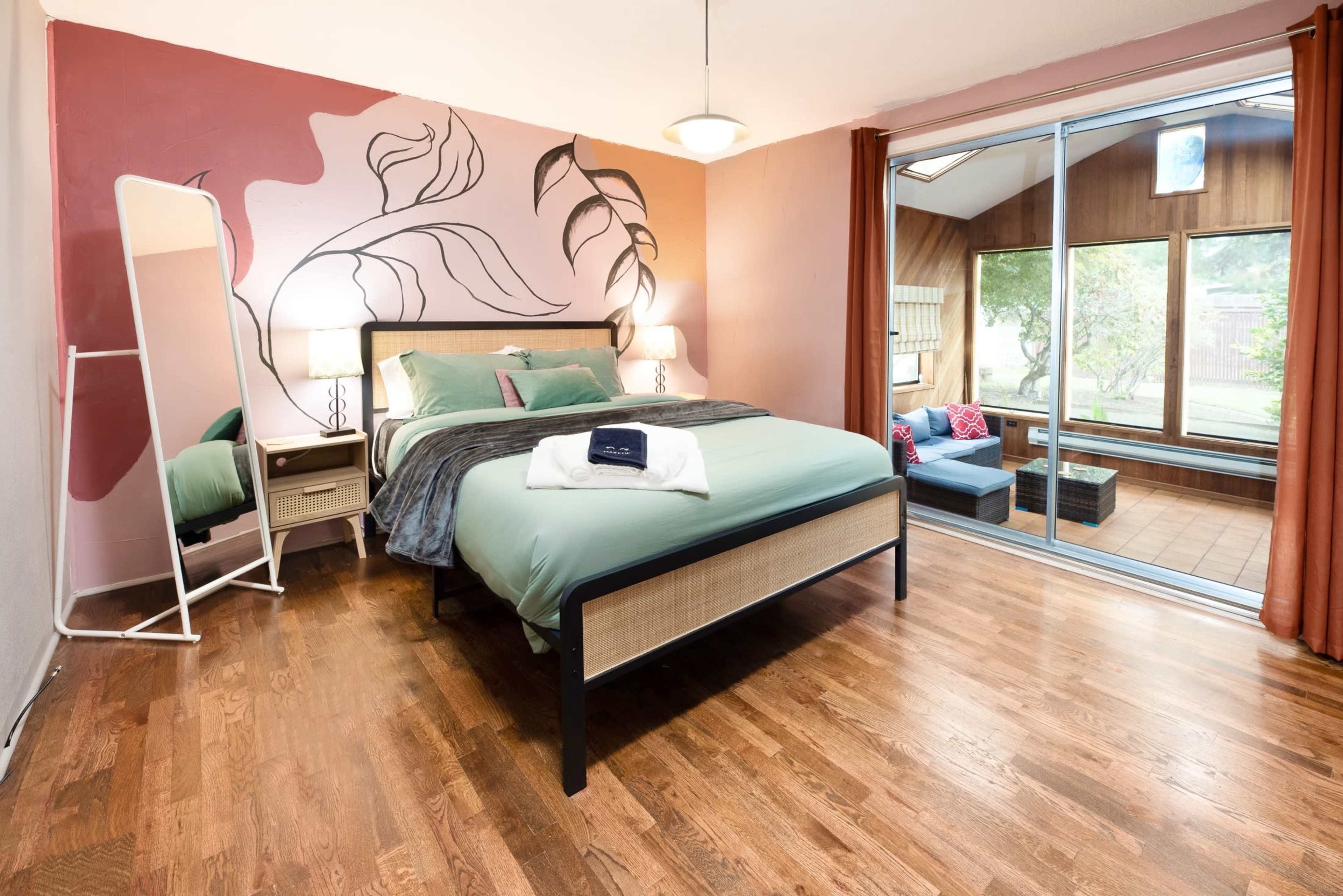 The room features a bed with teal bedding, a large mirror, wooden floors, and a wall painted with leafy designs, with a sunroom visible through large sliding glass doors.