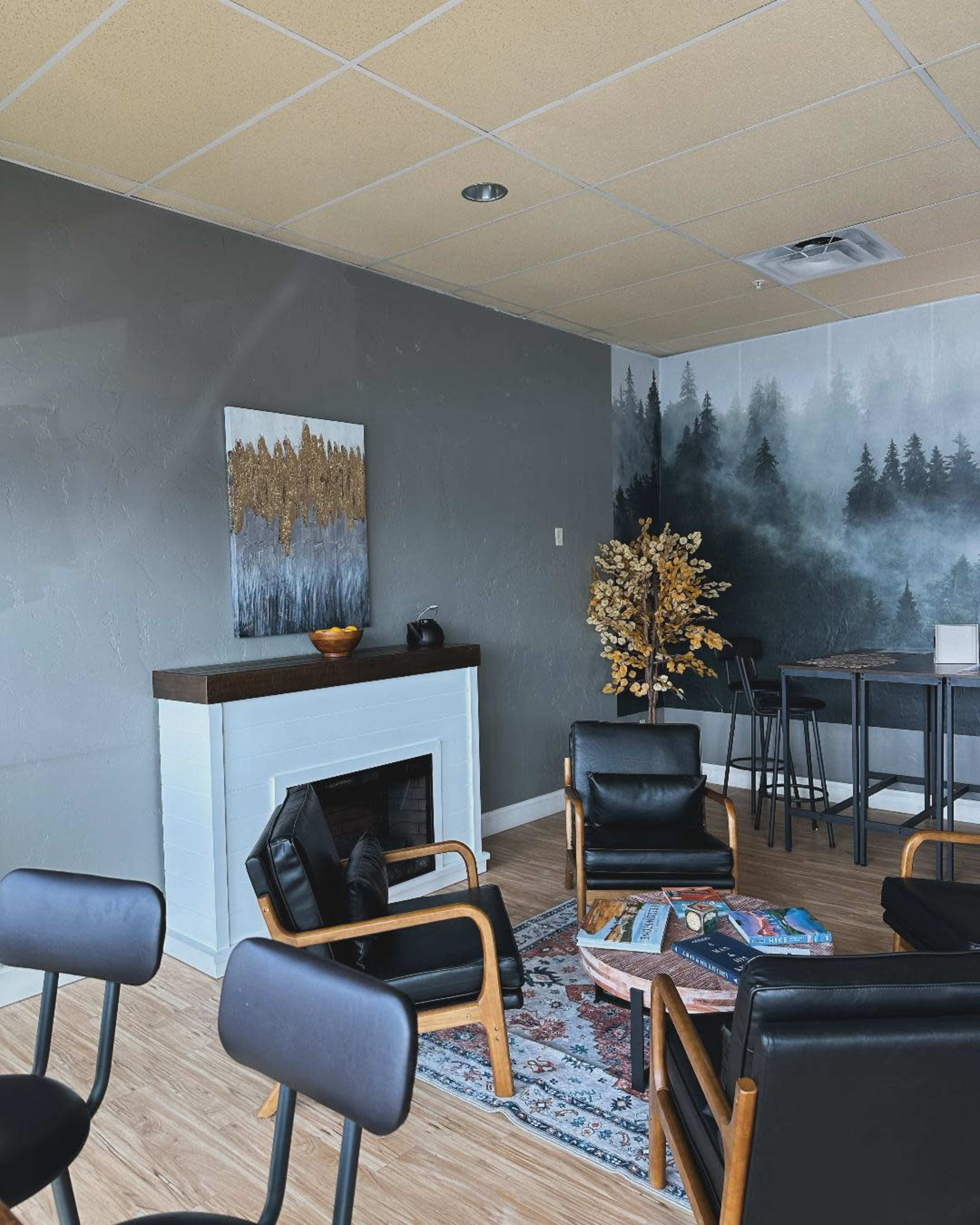 The image shows a cozy waiting area featuring a fireplace, black chairs, a table with decorative items, and a mural of a foggy forest on one wall.