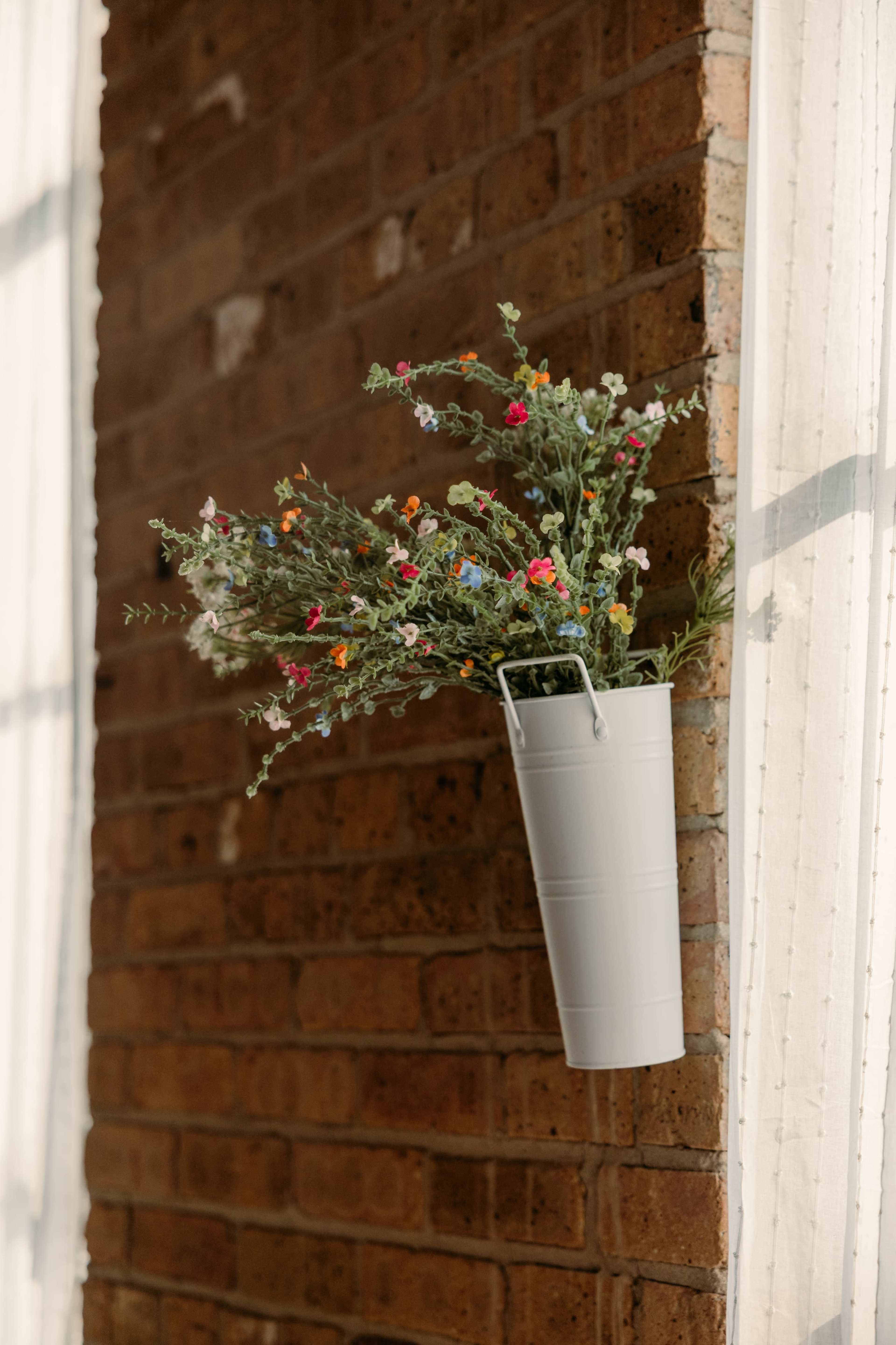 A white metal bucket filled with artificial flowers is mounted on a brick wall.