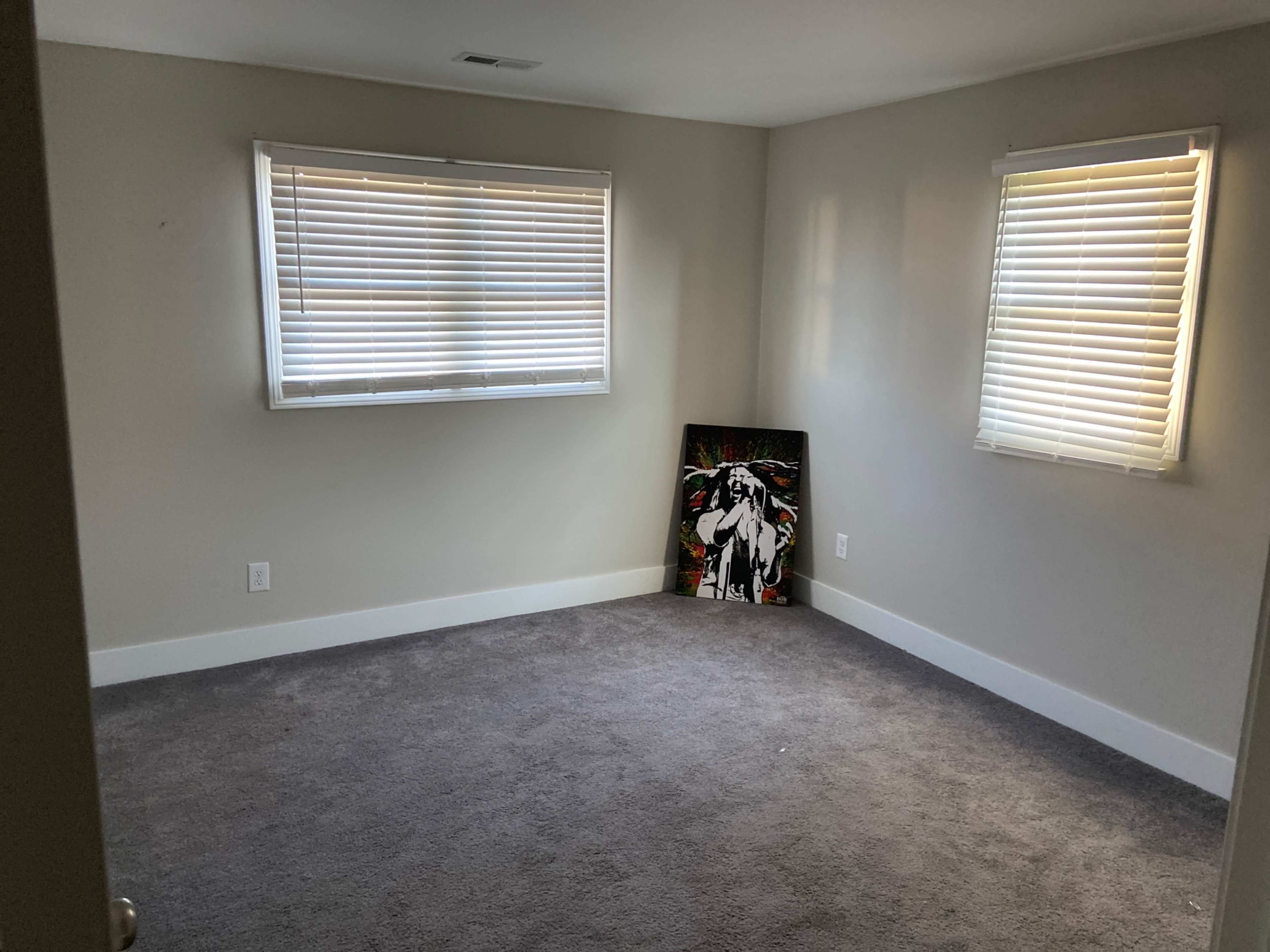 The room features gray carpet, two windows with blinds, and a colorful piece of artwork leaned against the wall.
