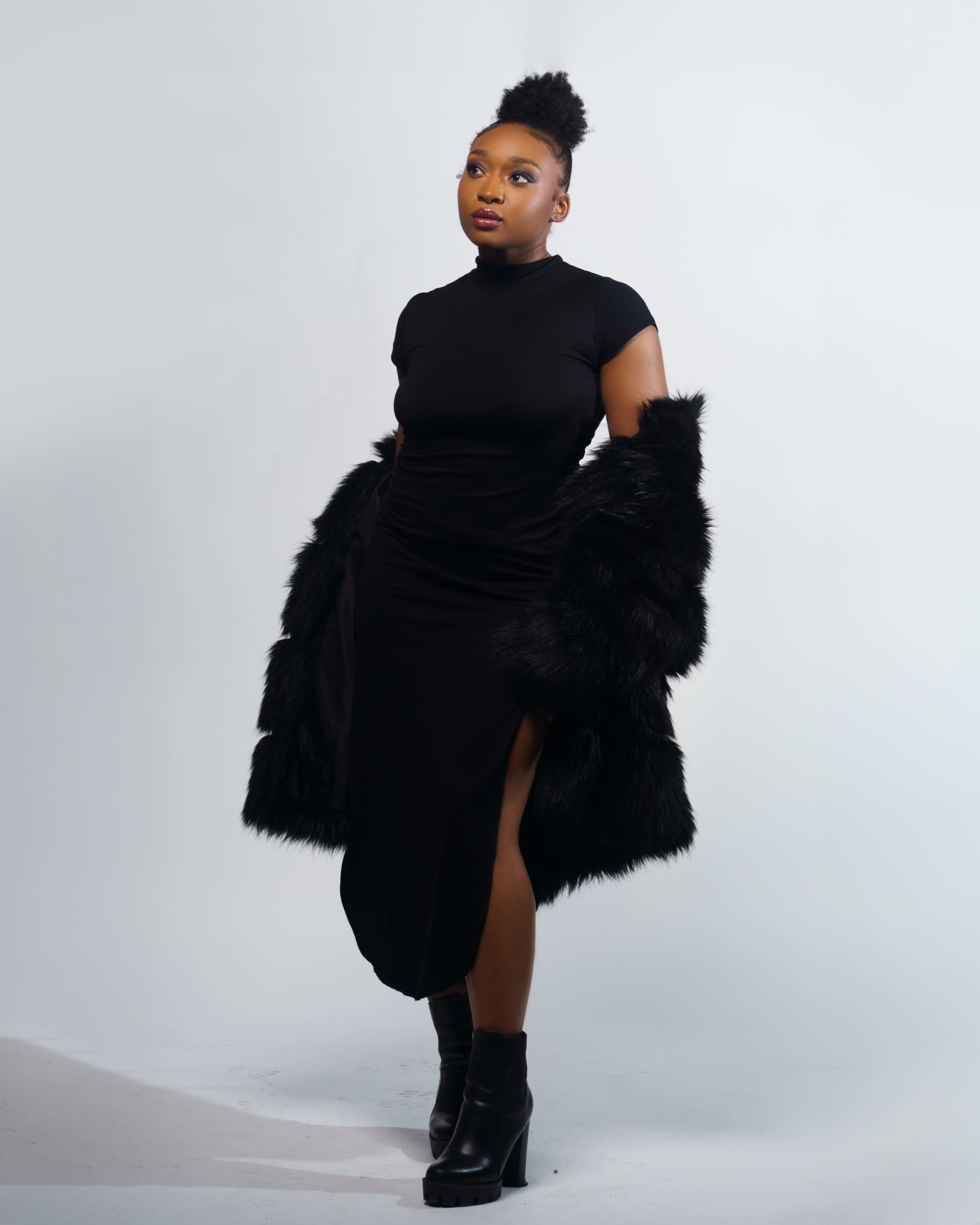 A woman poses in a black dress and fur coat against a plain background.