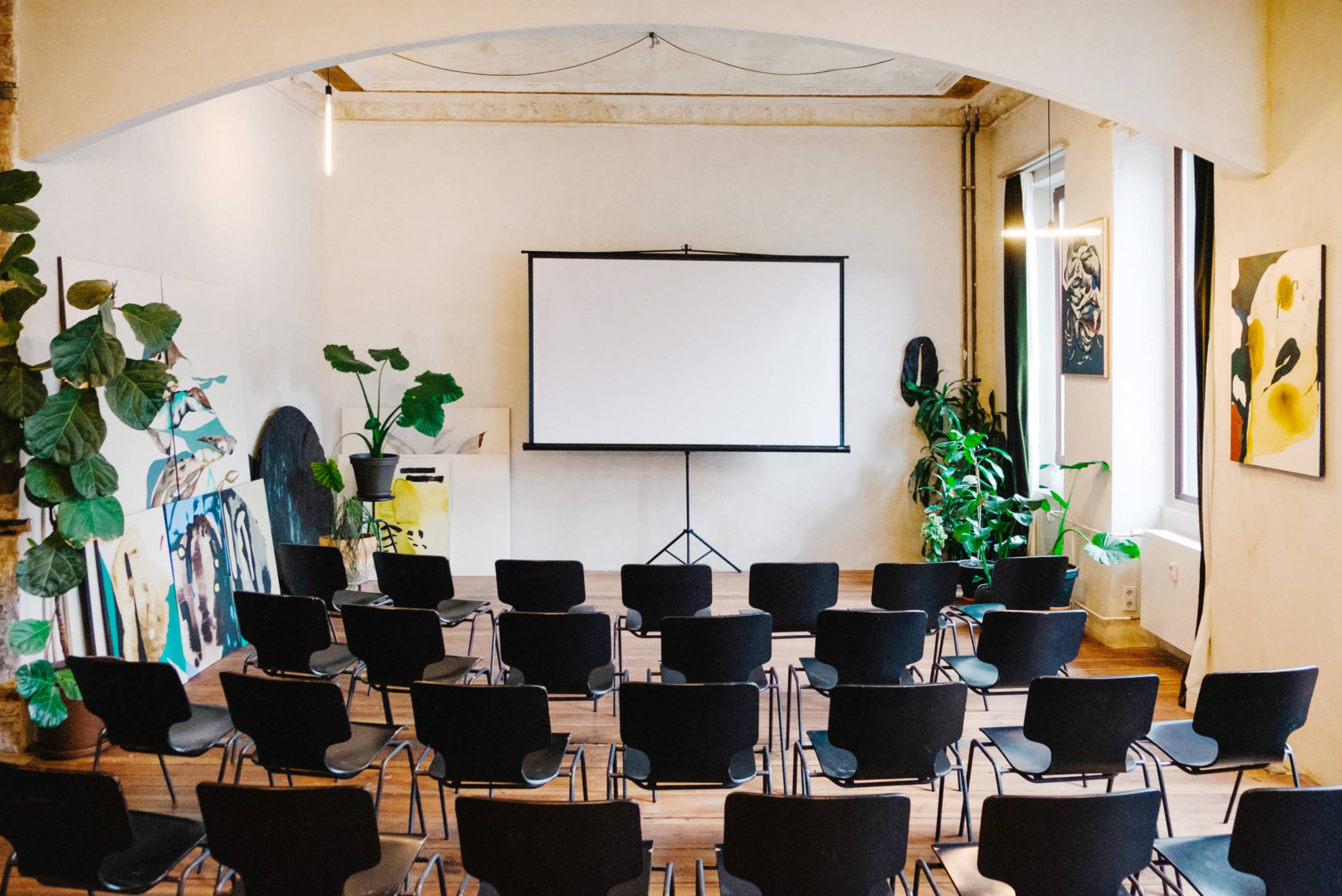 A spacious room features neatly arranged black chairs facing a large projection screen, surrounded by plants and artwork on the walls.