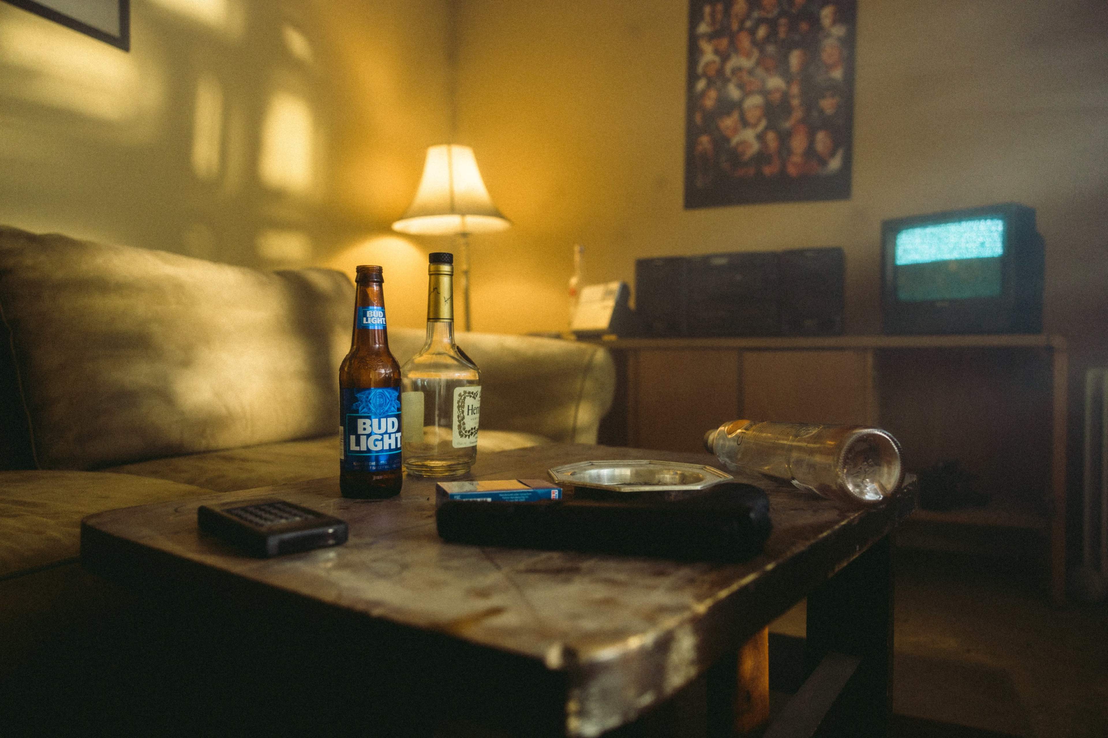 A small living room scene features a worn wooden coffee table with two bottles of beer, a liquor bottle, an empty glass, and a remote control, illuminated by a lamp in the background.