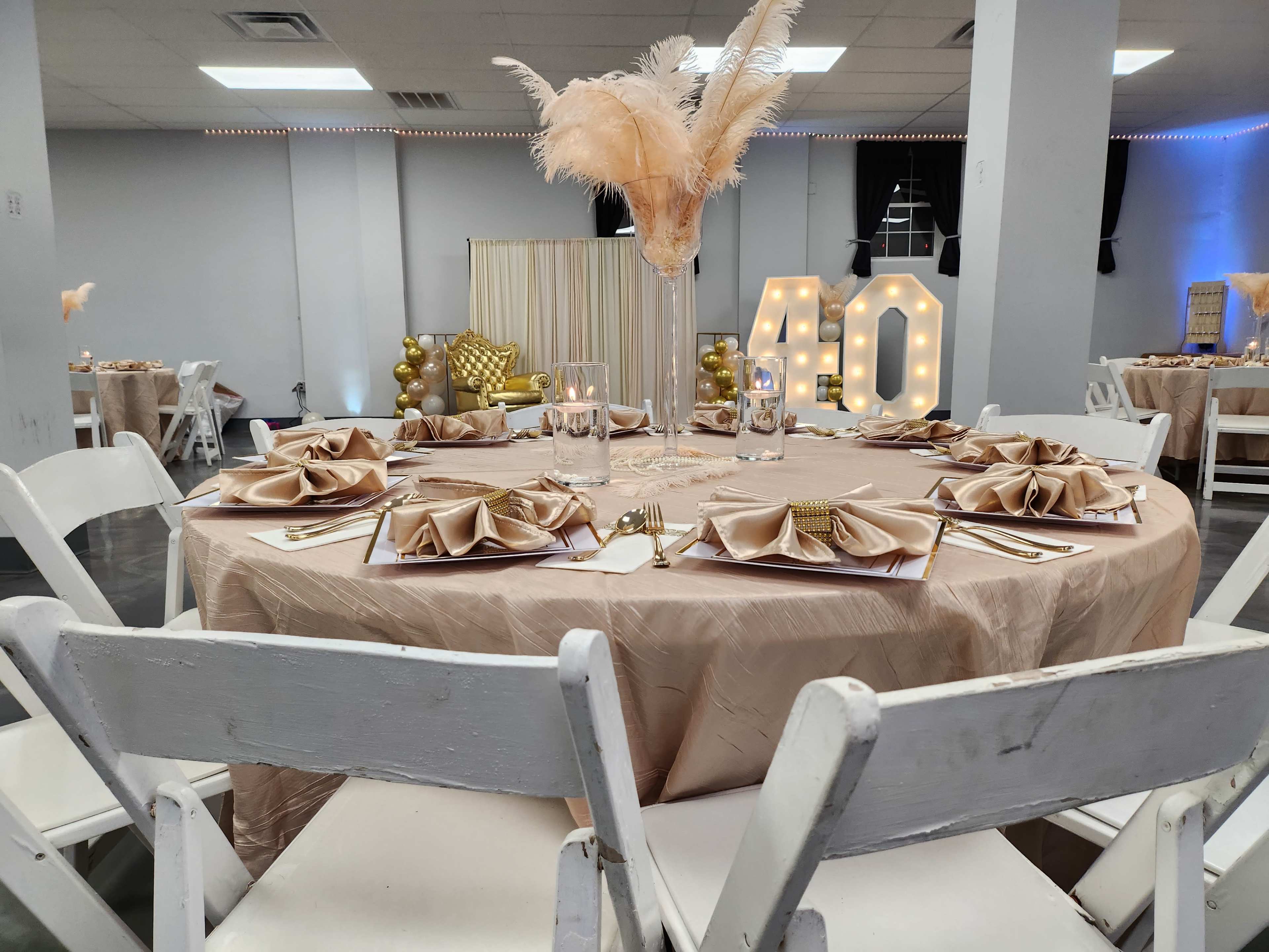 The image shows a banquet hall set up for a celebration, featuring round tables dressed in beige tablecloths, elegant place settings, and a large illuminated "40" display in the background.