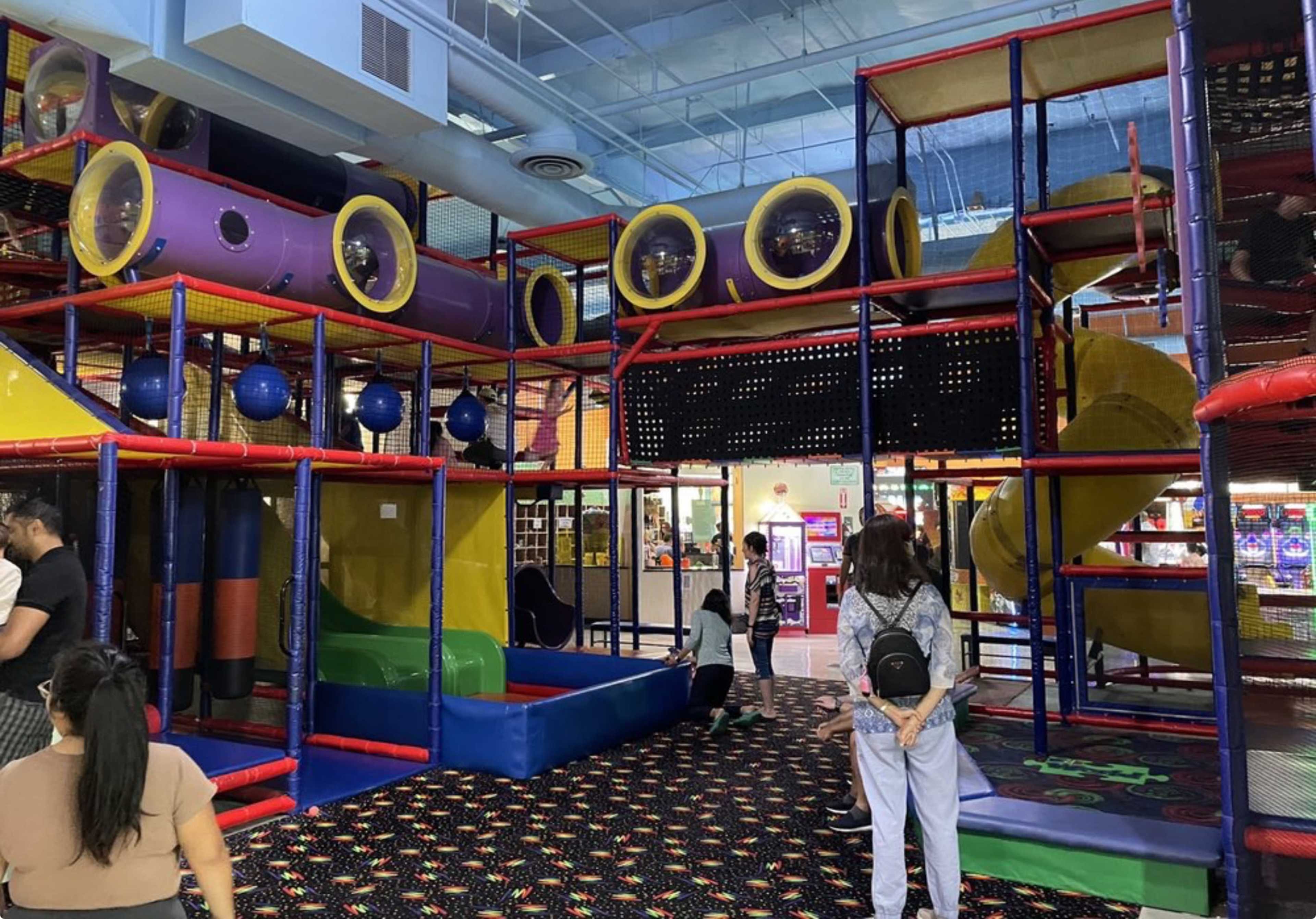 The image shows an indoor play area with colorful structures, including climbing tunnels, slides, and soft play features, surrounded by families and children.