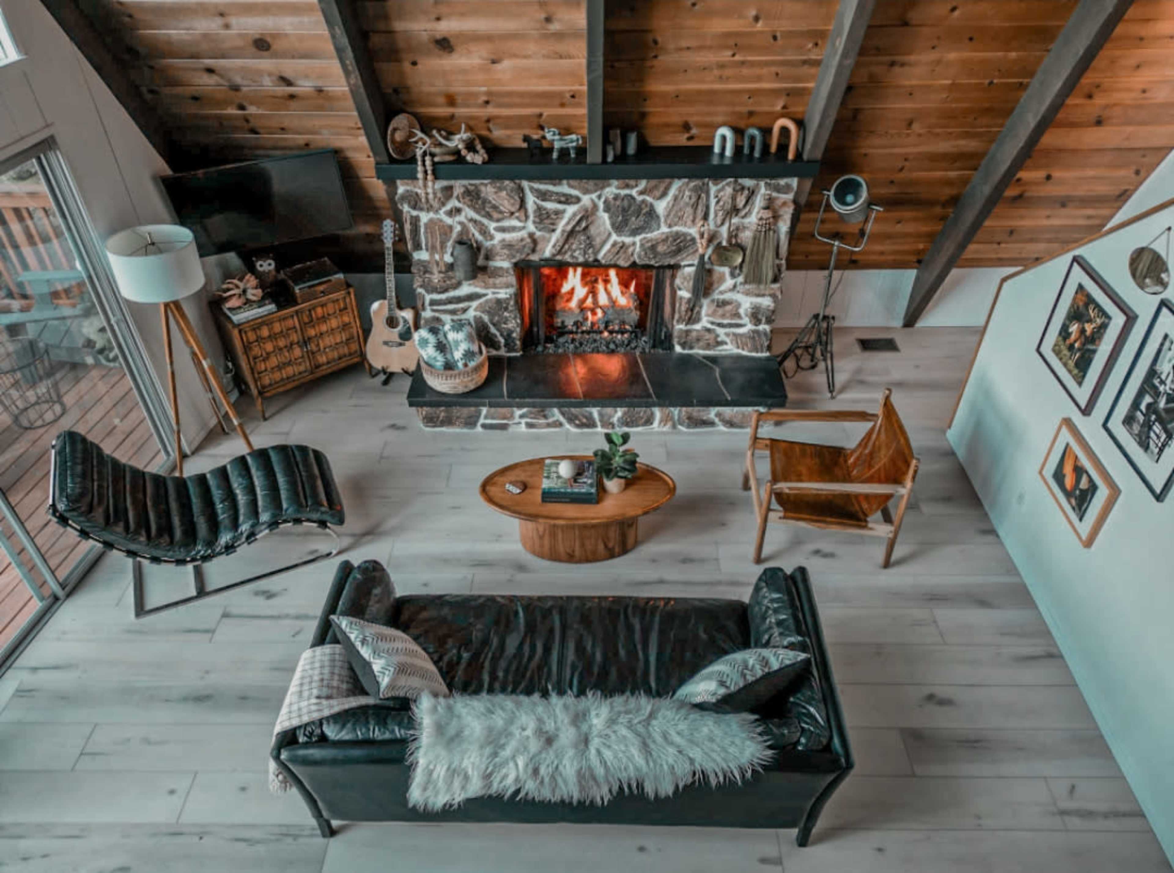 A cozy living room features a stone fireplace, wooden beams, a black leather sofa, a wooden coffee table, and various seating arrangements.