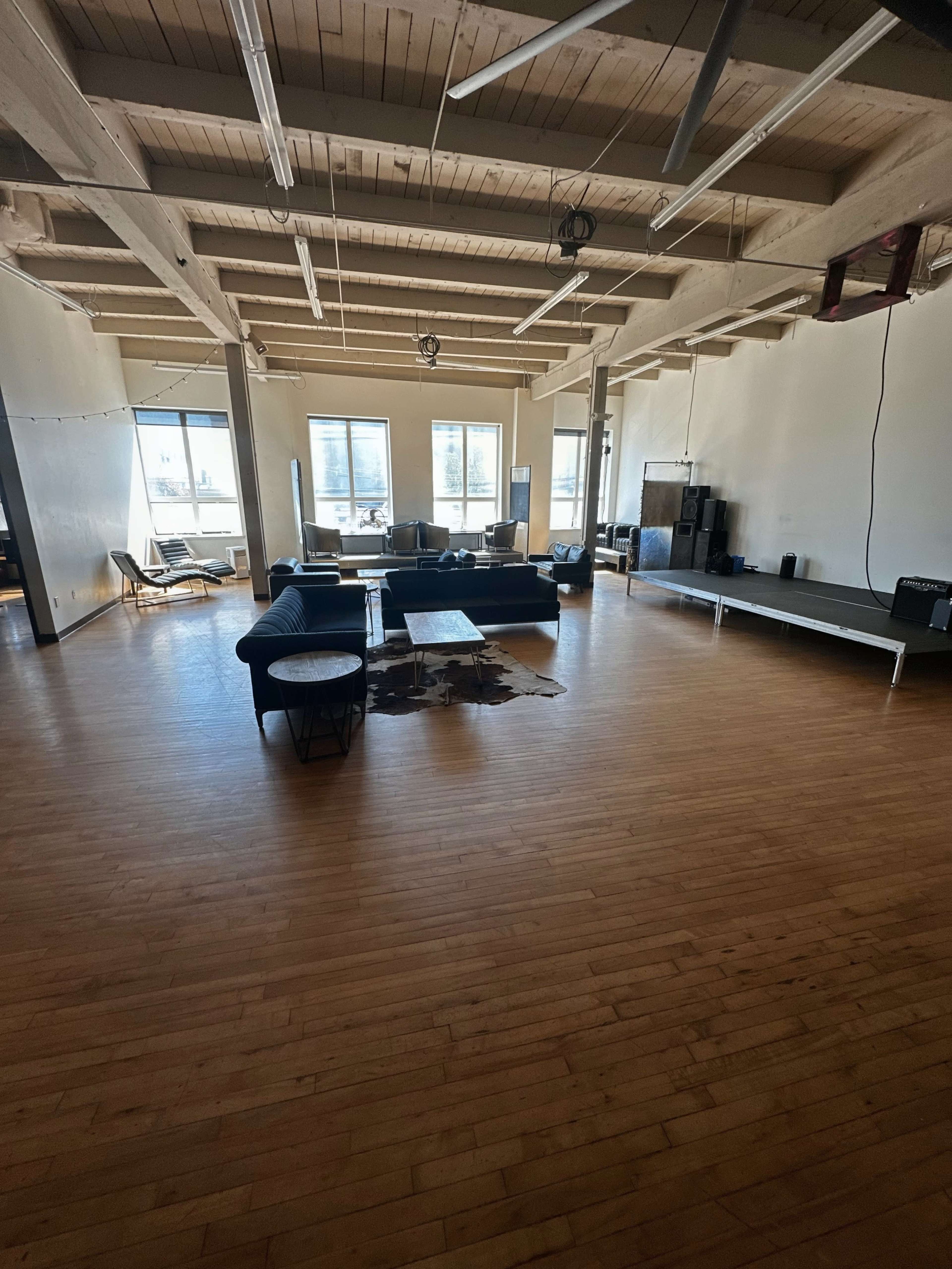 The image shows a spacious, open living area with wooden floors, several large windows, and minimalist furniture arrangements.