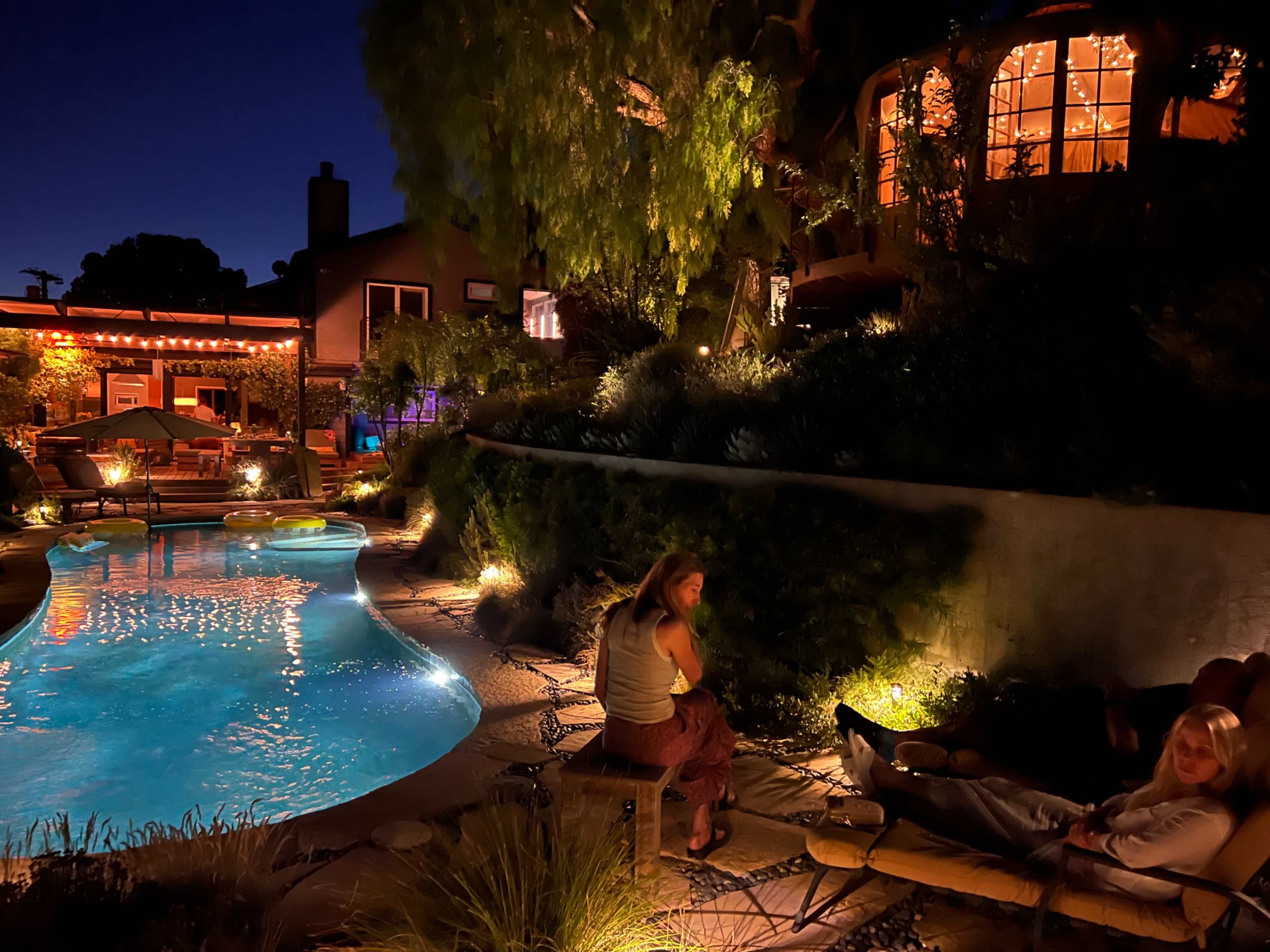 The scene features a well-lit outdoor area with a pool surrounded by seating and lush greenery, illuminated by soft lights at night.