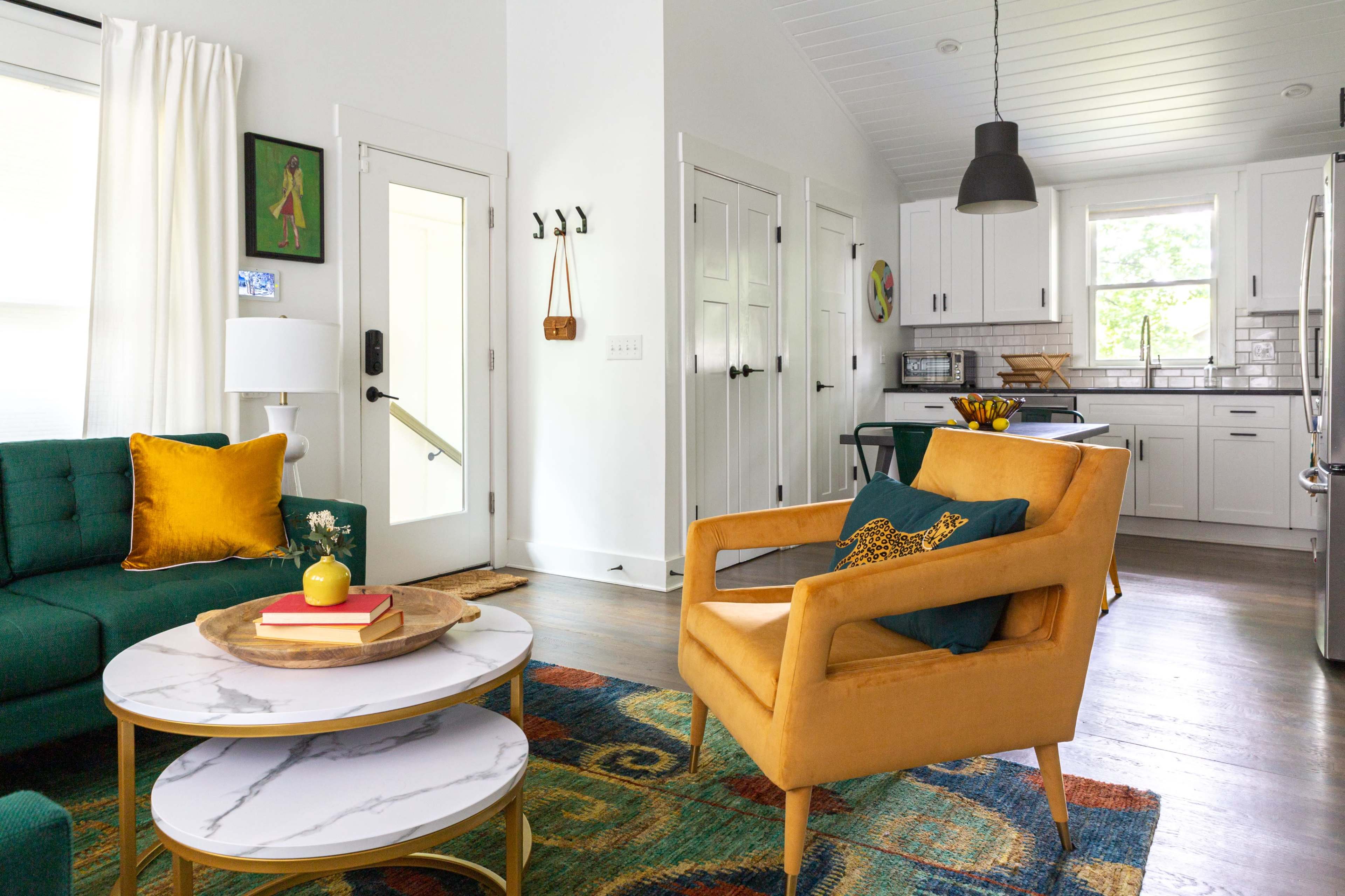 A modern living room with a green sofa, a yellow armchair, and a marble-top coffee table, adjacent to a kitchen with white cabinetry and natural light.