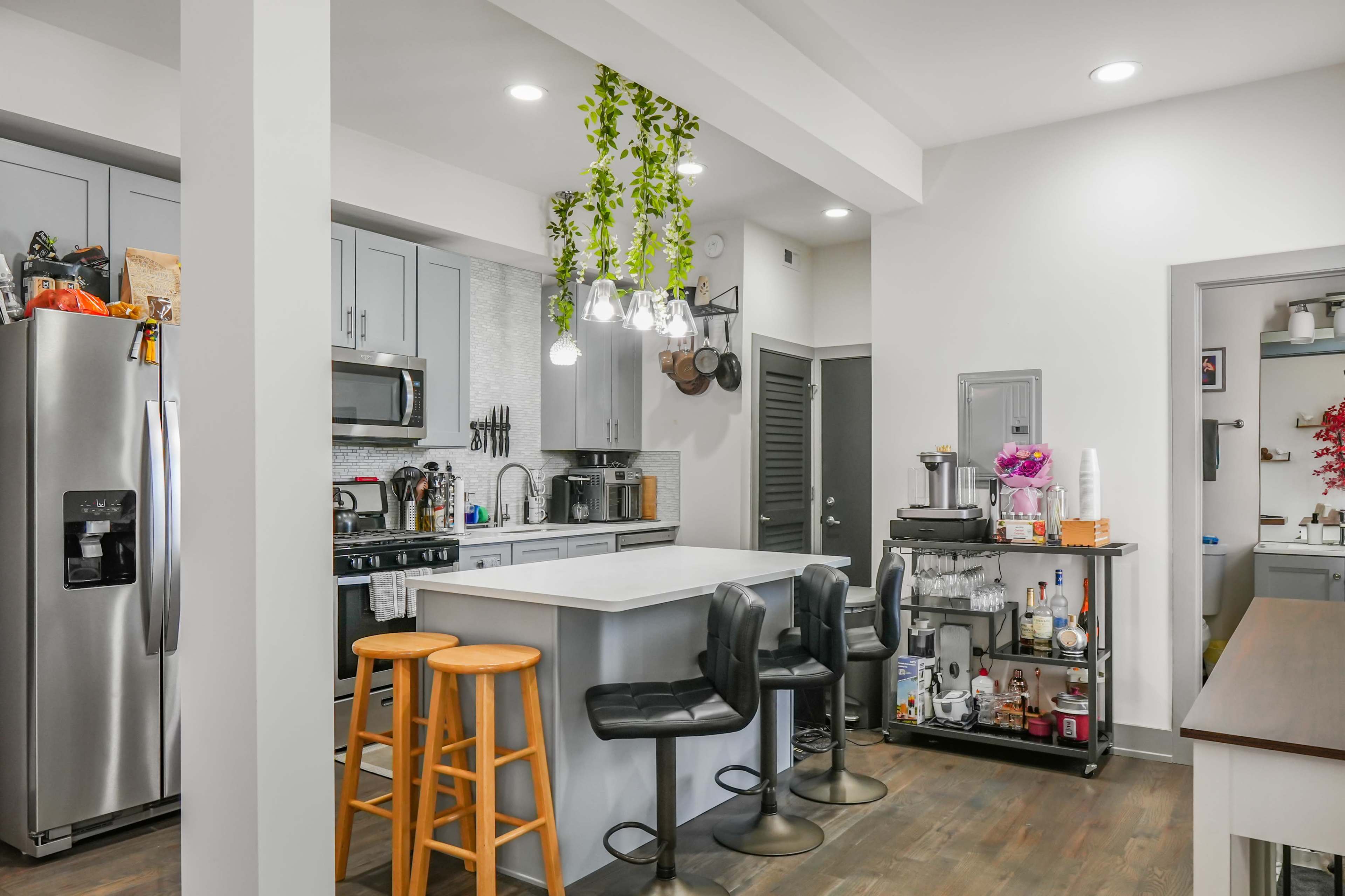 The image shows a modern kitchen featuring stainless steel appliances, a large island with seating, and a mix of bar stools and chairs, along with decorative plants hanging from the ceiling.