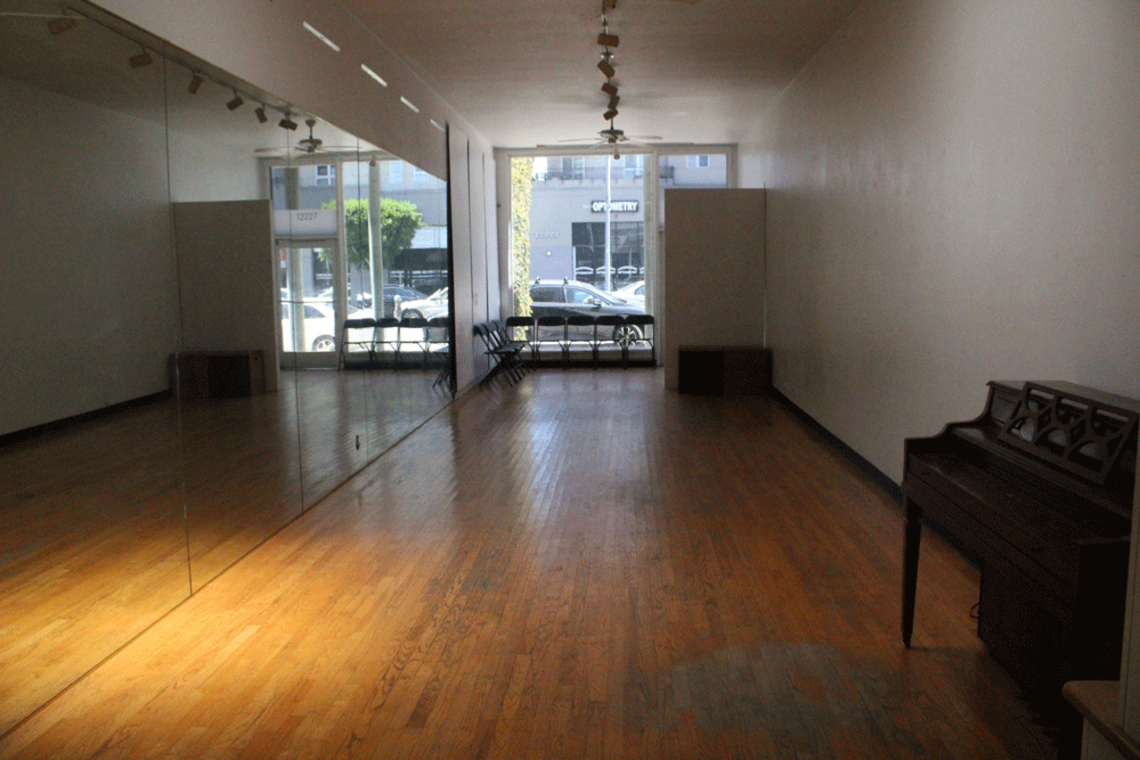 The image shows a spacious room with wooden flooring, large mirrors on one side, and natural light coming in through a window.