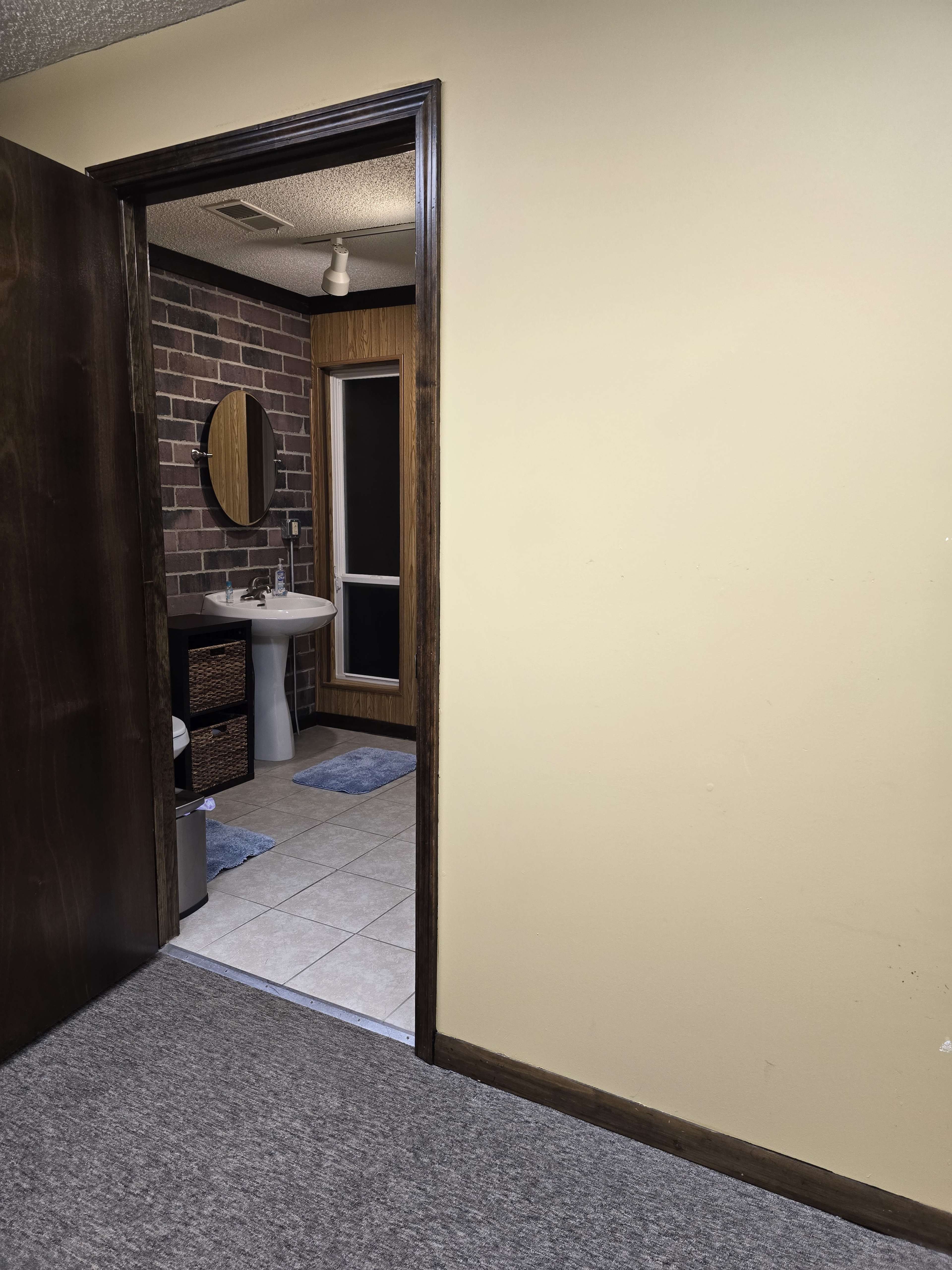 An open door leading to a bathroom with a brick wall, a sink, a mirror, and a door to a shower.