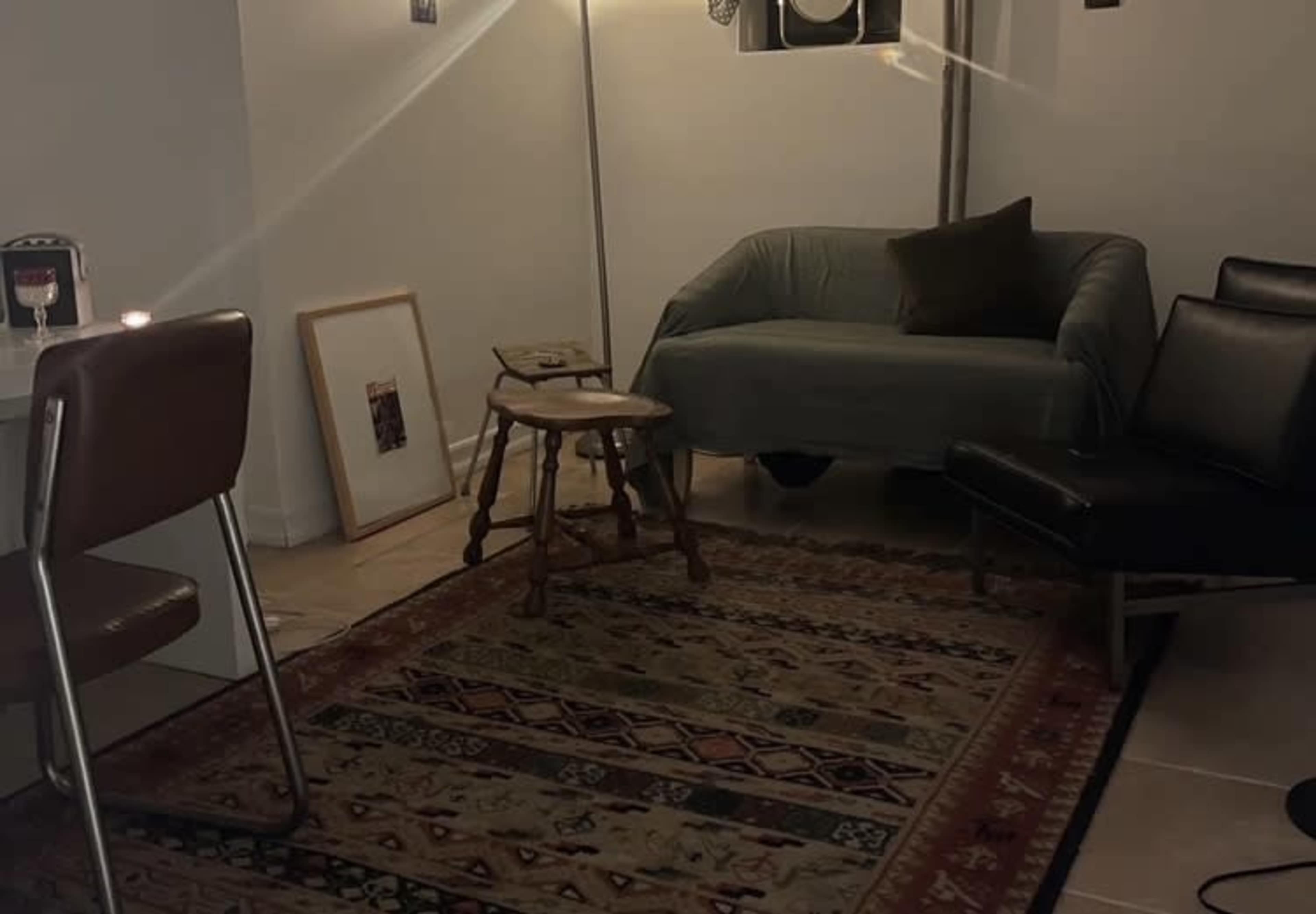 The image shows a cozy room with a green couch, a wooden stool, and a black leather chair, all arranged on a patterned rug.