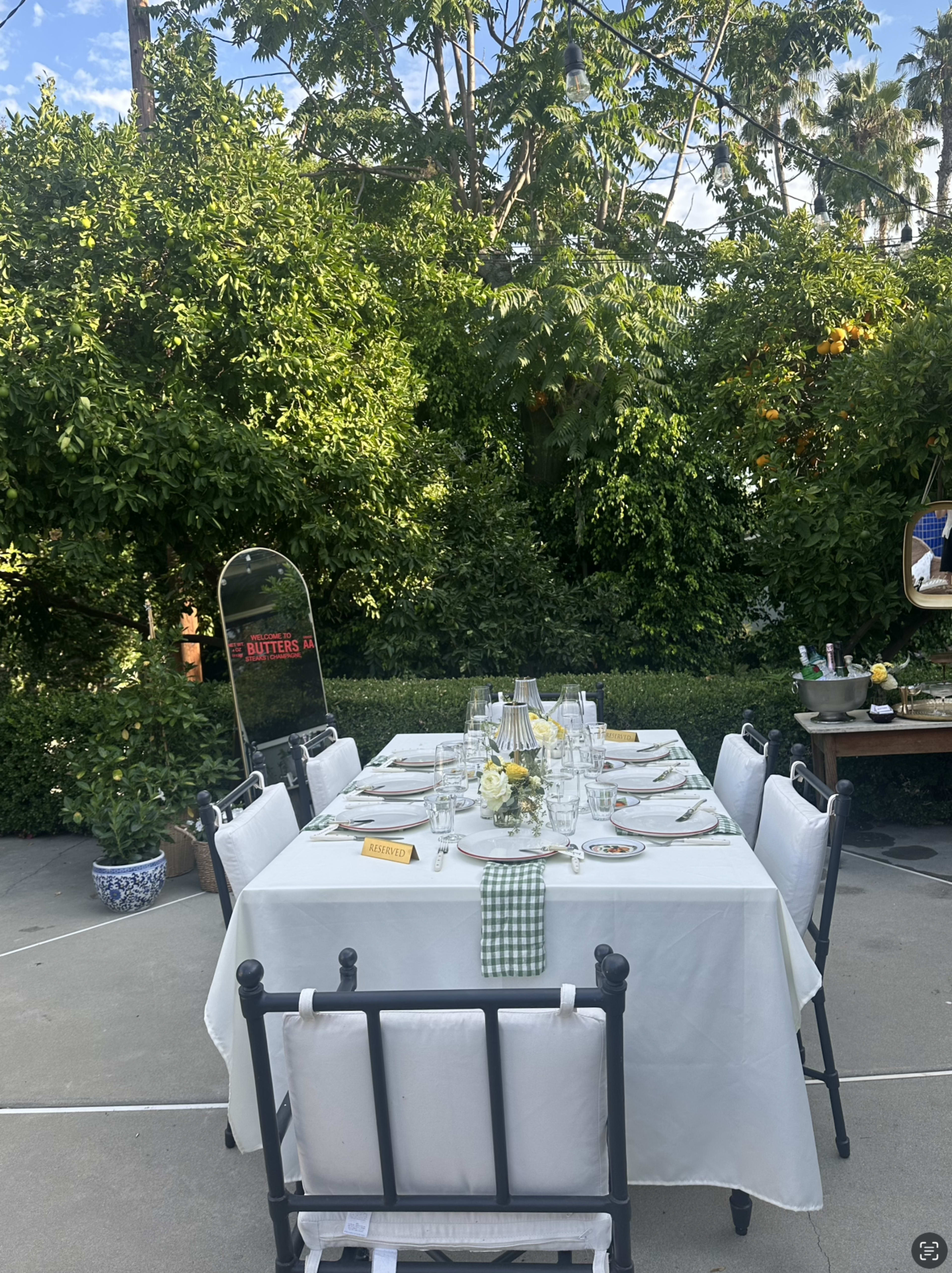 A large outdoor dining table is set with plates, utensils, and a floral centerpiece, surrounded by lush greenery.