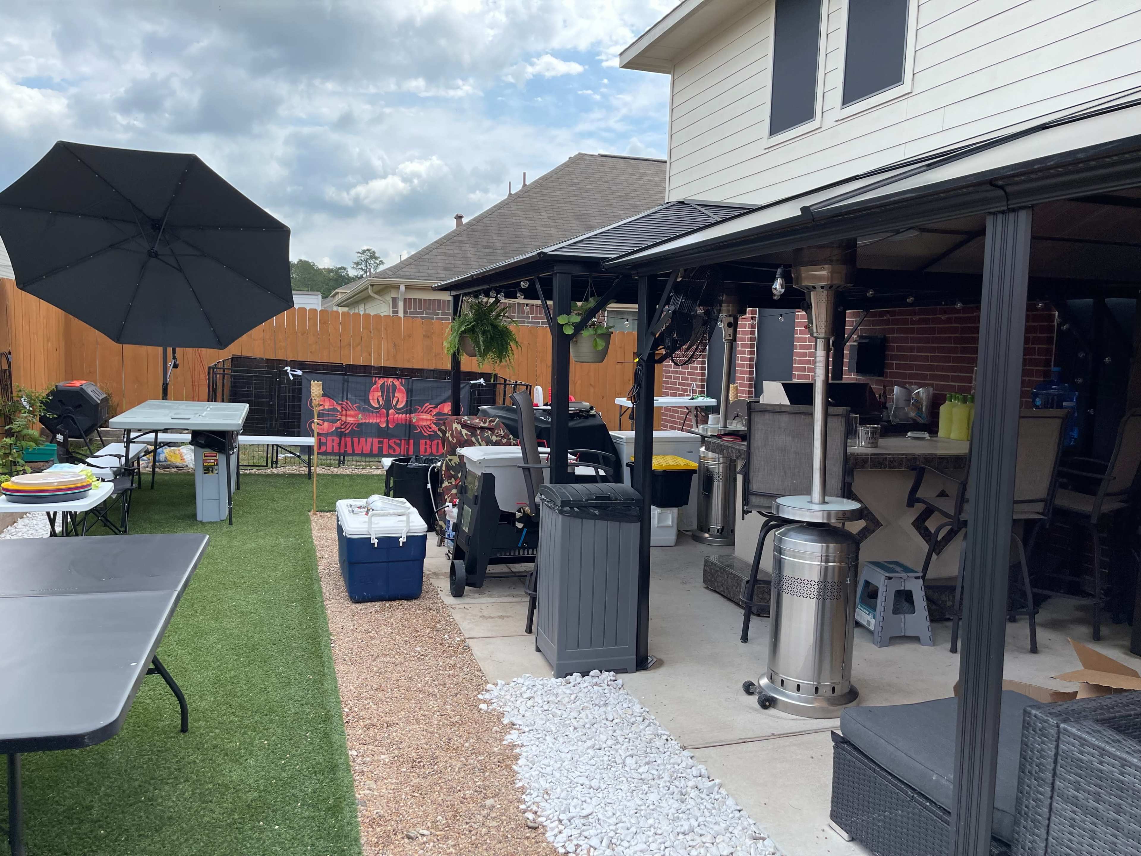 The image shows a backyard setup featuring a gravel and grass area with tables, a BBQ grill, a cooler, and an umbrella beside a large outdoor cooking space, all enclosed by a wooden fence.