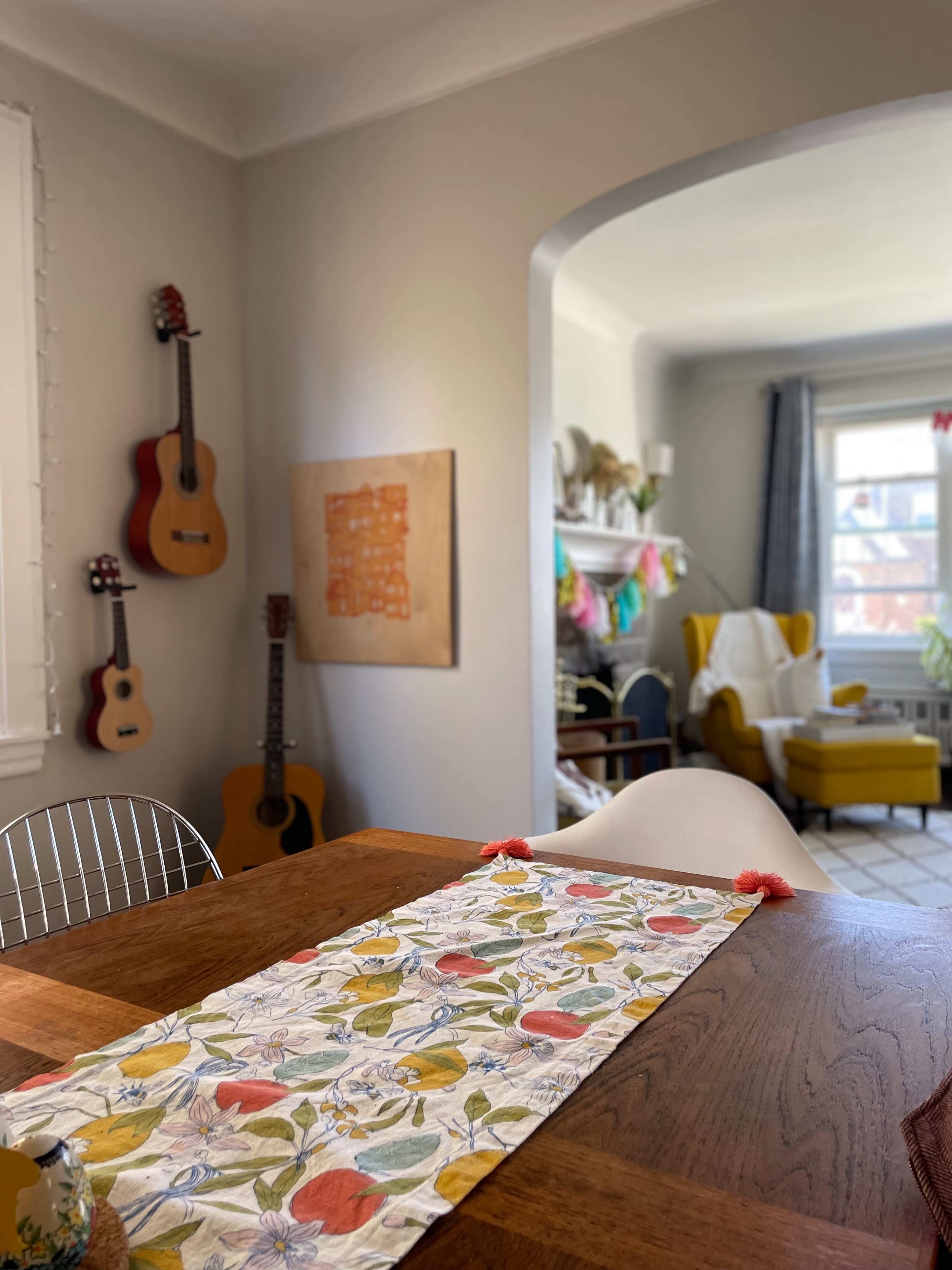 A dining table with a colorful table runner is in the foreground, while guitars are mounted on the wall and a bright armchair is visible in the background of a well-lit room.