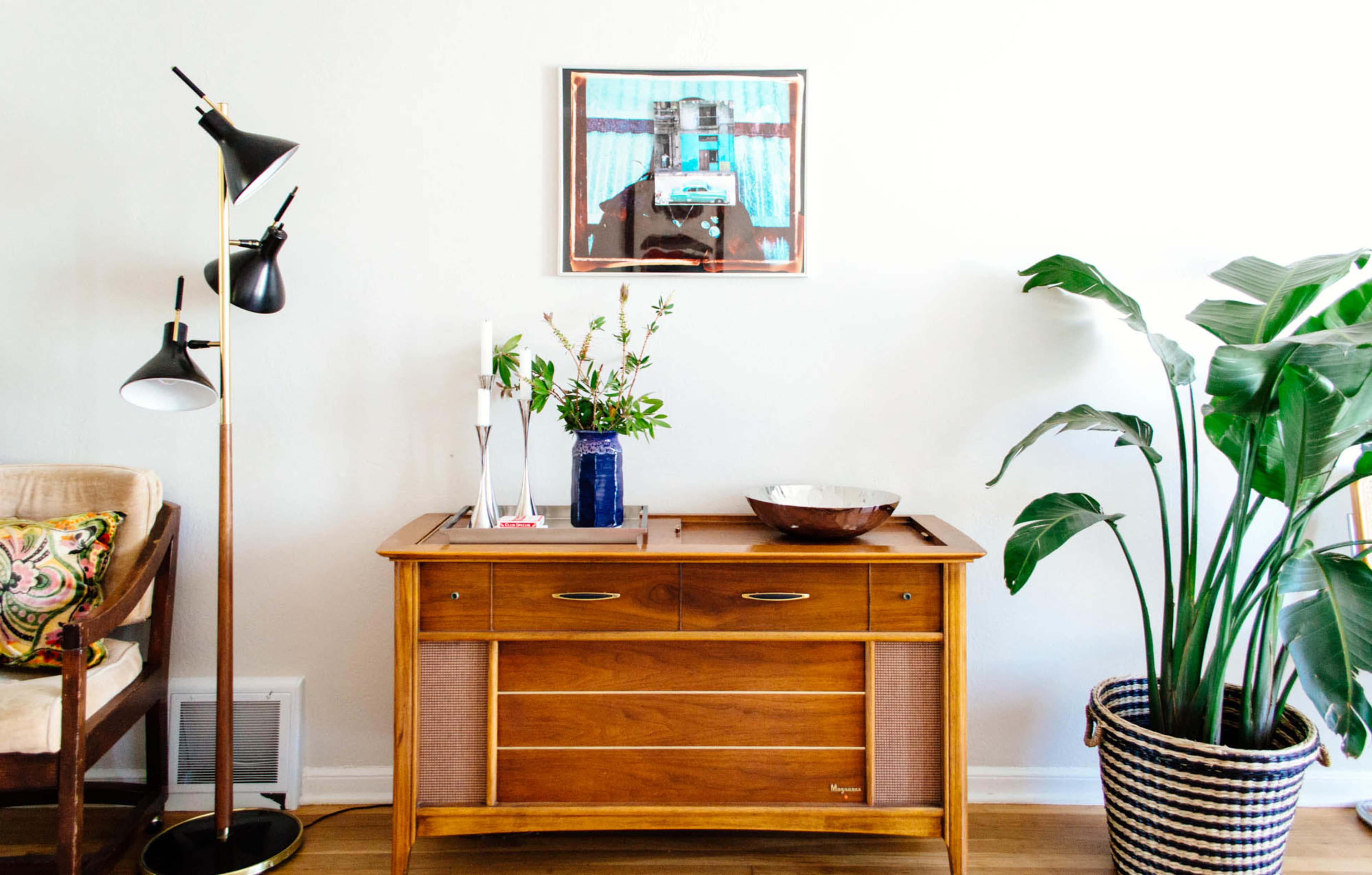 A wooden sideboard with a decorative bowl and a plant sits beneath a framed artwork on a light-colored wall, accompanied by a black floor lamp and a chair in a corner.