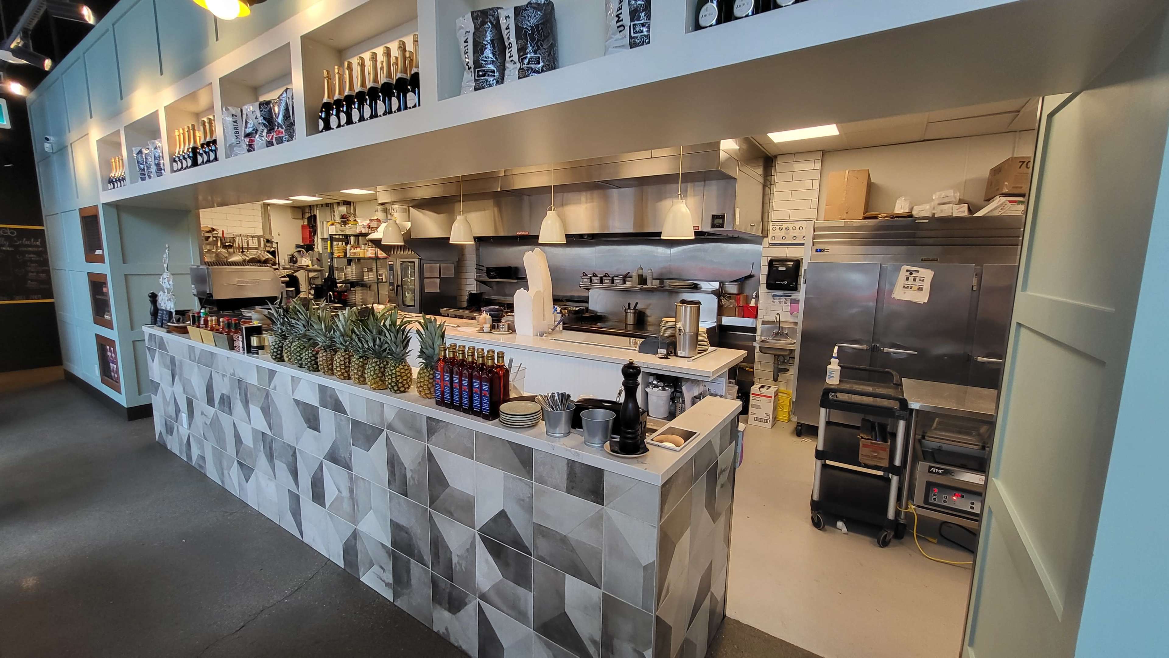 The image shows a modern café or restaurant interior, featuring a stylish counter with a geometric design, a display of pineapples, coffee equipment, and a visible kitchen area in the background.