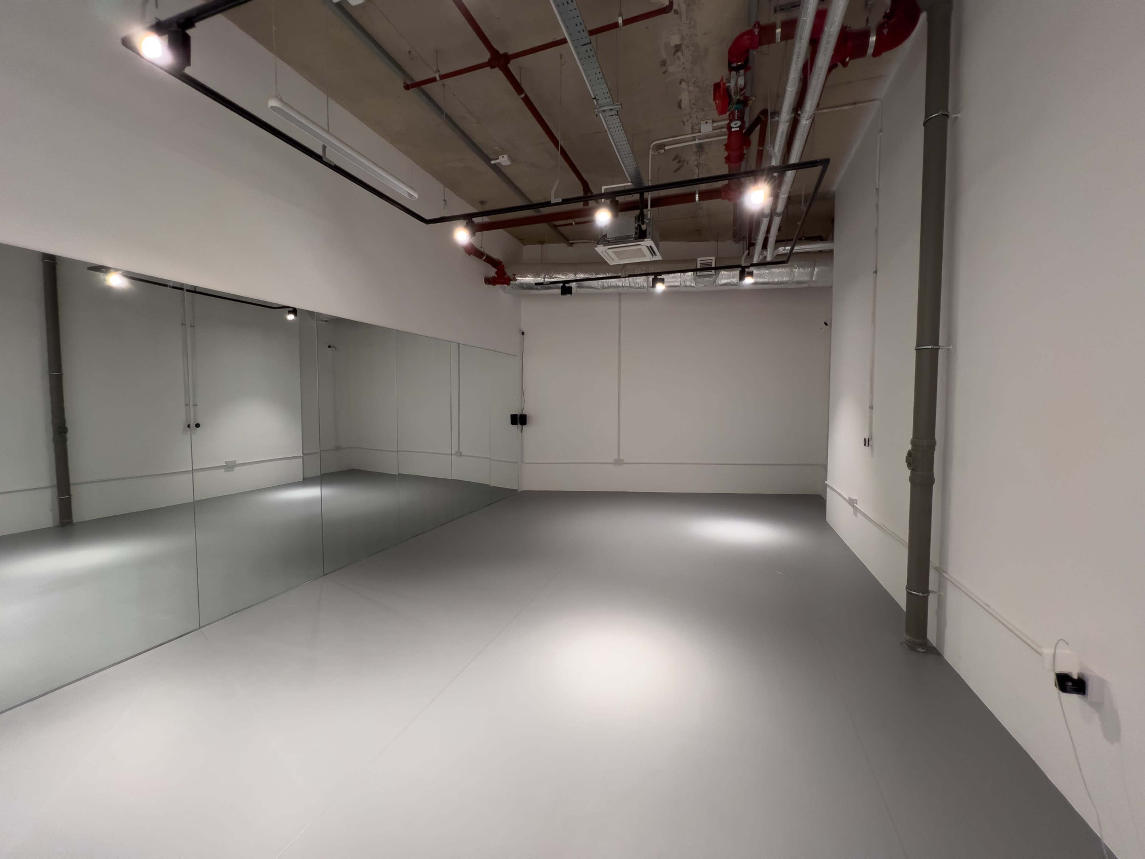 A spacious room with gray flooring and a large mirror on one wall, illuminated by overhead lights.