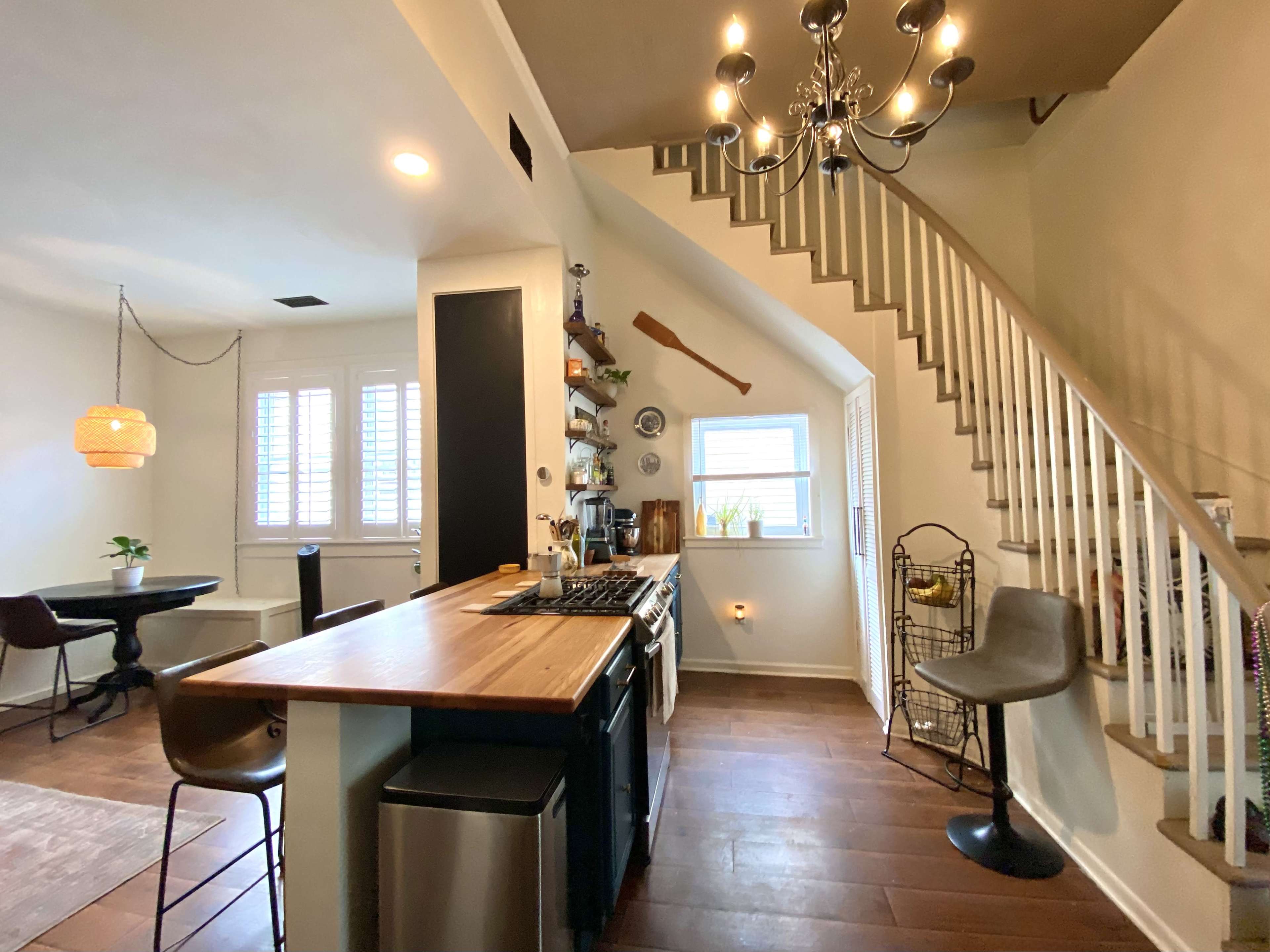 A modern kitchen features a wooden countertop, stainless steel appliances, and an adjacent dining area with a staircase and decorative chandelier.