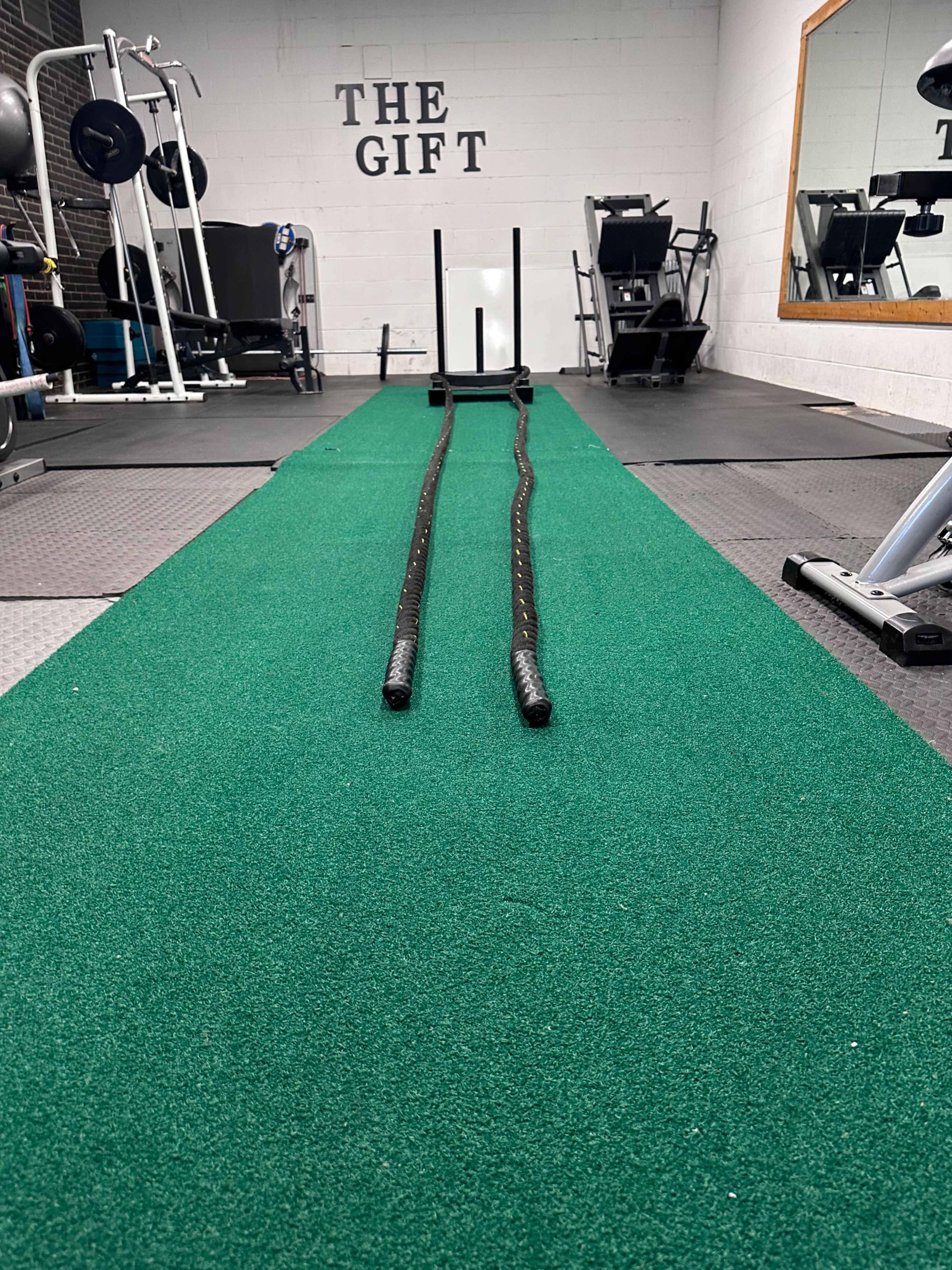 A spacious gym features a green turf area with two battle ropes laid out, leading towards workout equipment in the background.