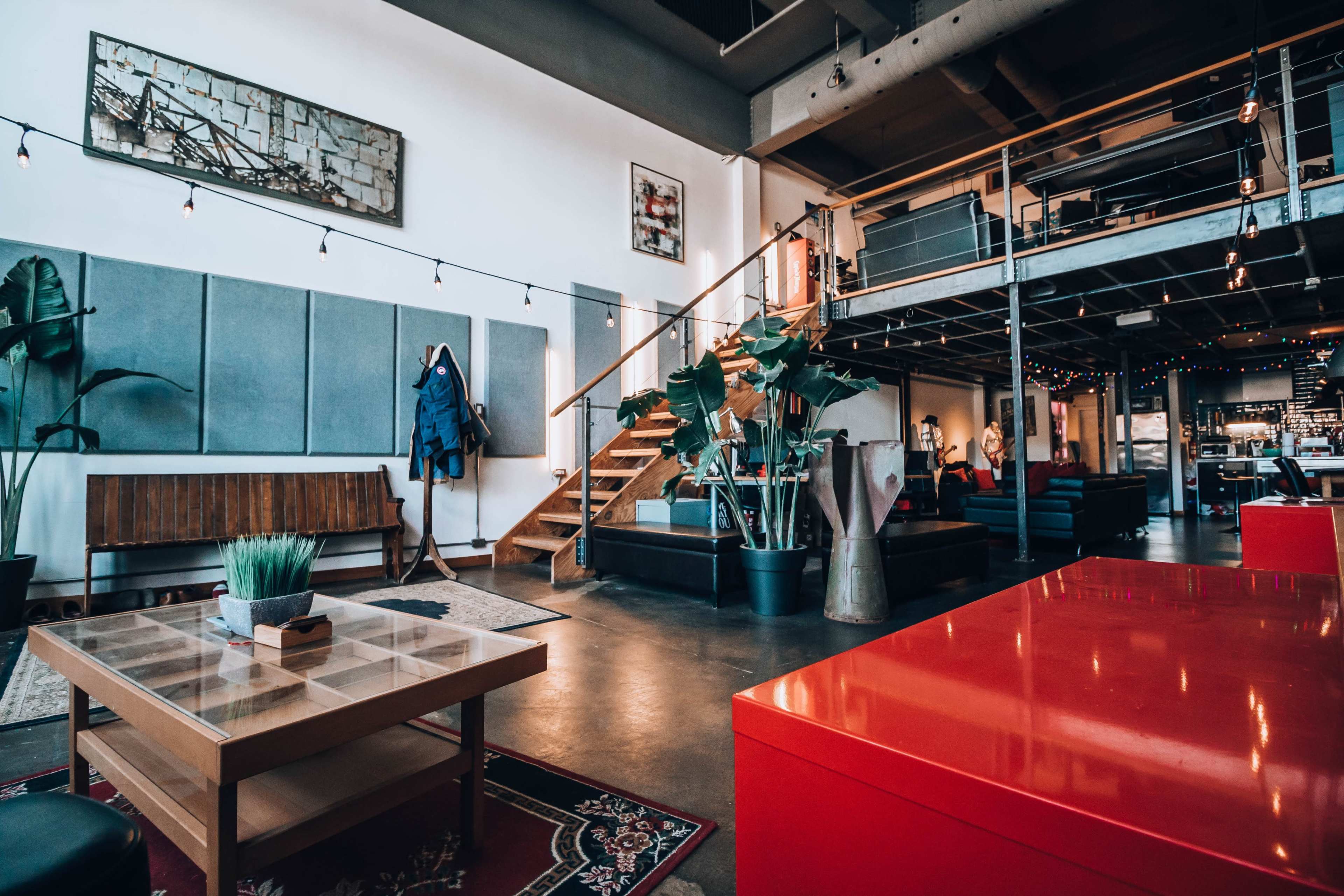 A modern, open-concept living space with wooden stairs, plants, and a mix of furniture, including a red table and a couch, illuminated by string lights.