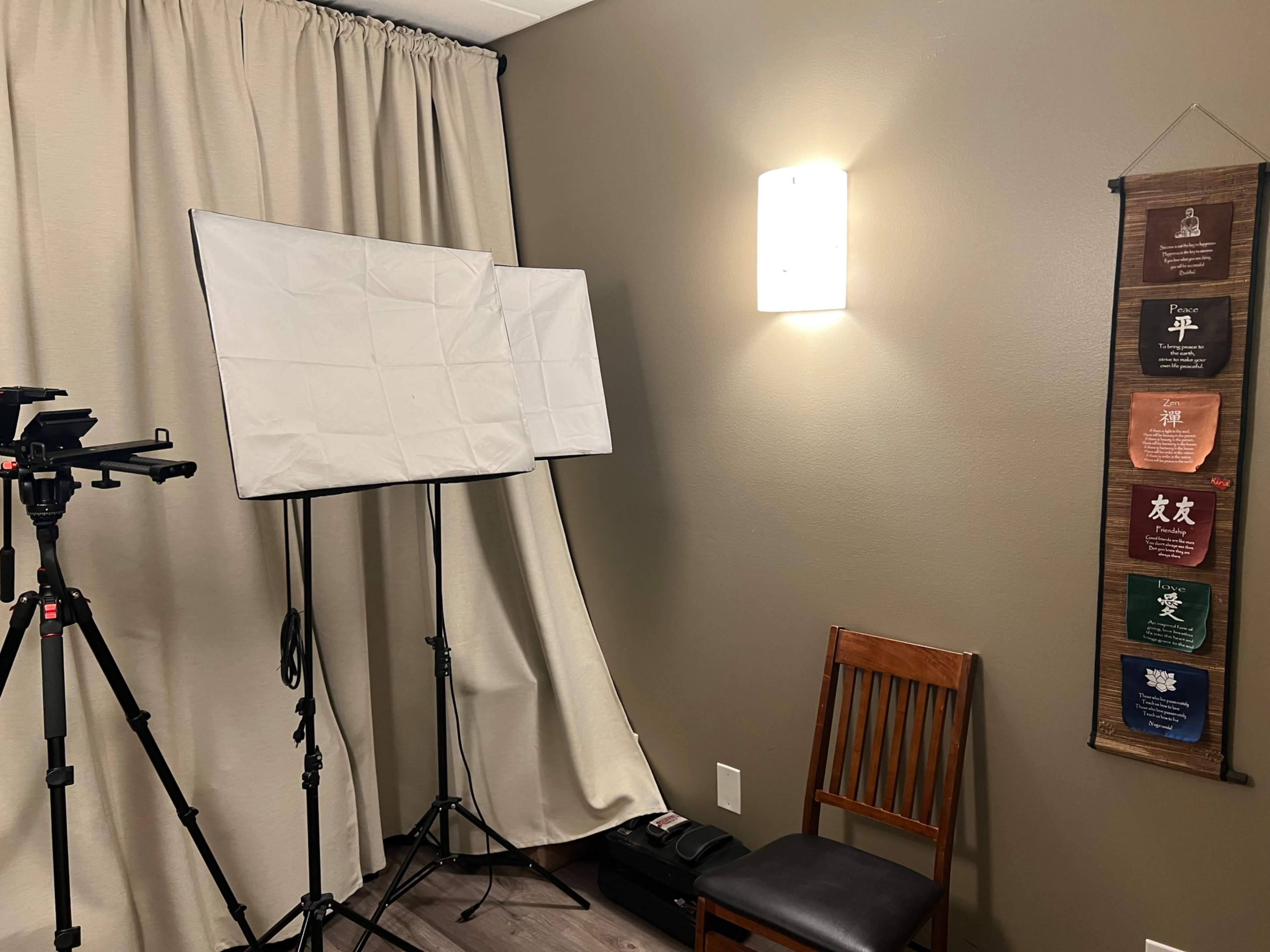 The image shows a small room with a chair, a camera setup on a tripod, softbox lights, and a wall hanging with various colored panels.