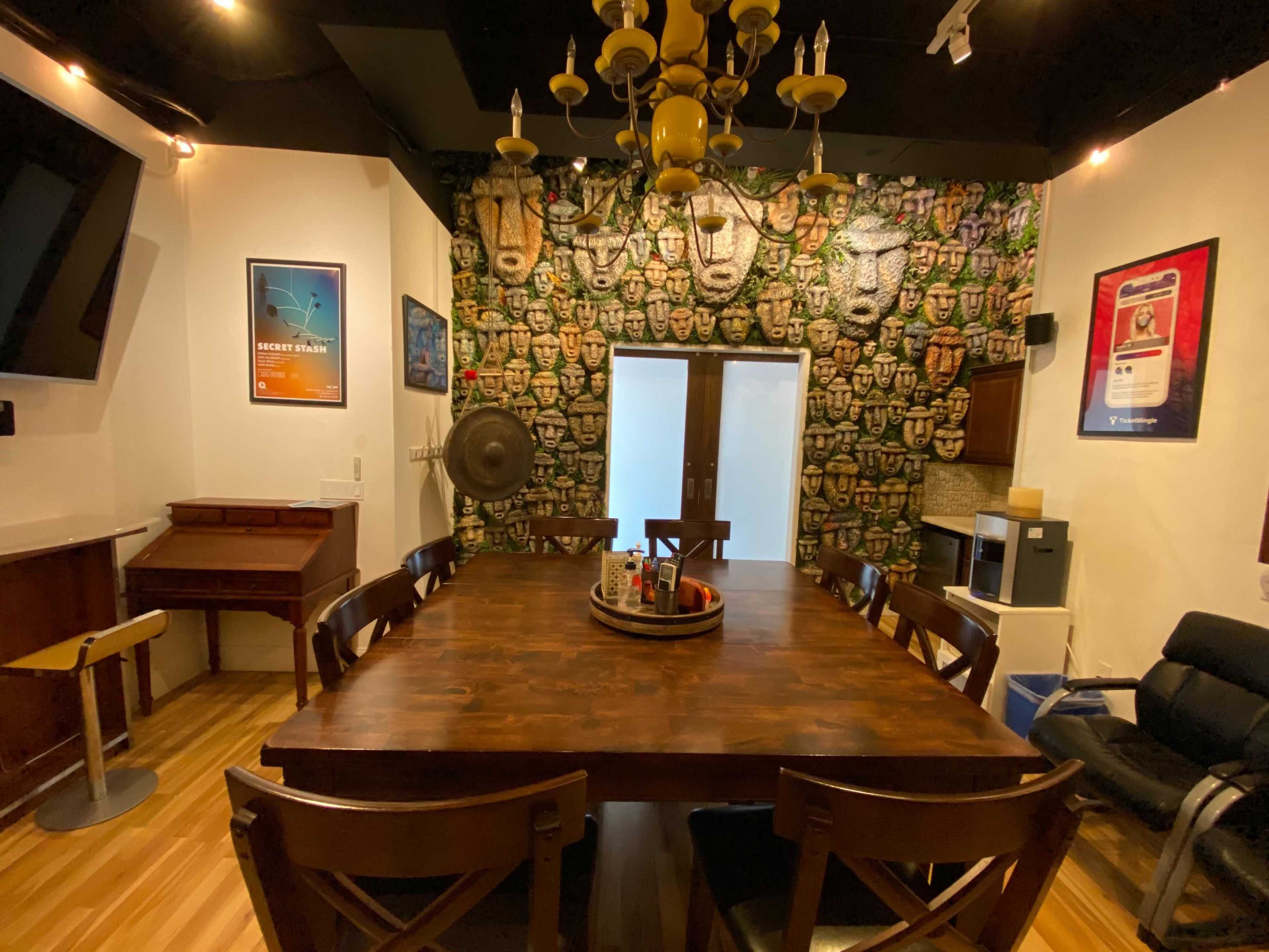 A dining area featuring a large wooden table surrounded by chairs, a wall adorned with patterned masks, and various furnishings including a piano and refrigerator.