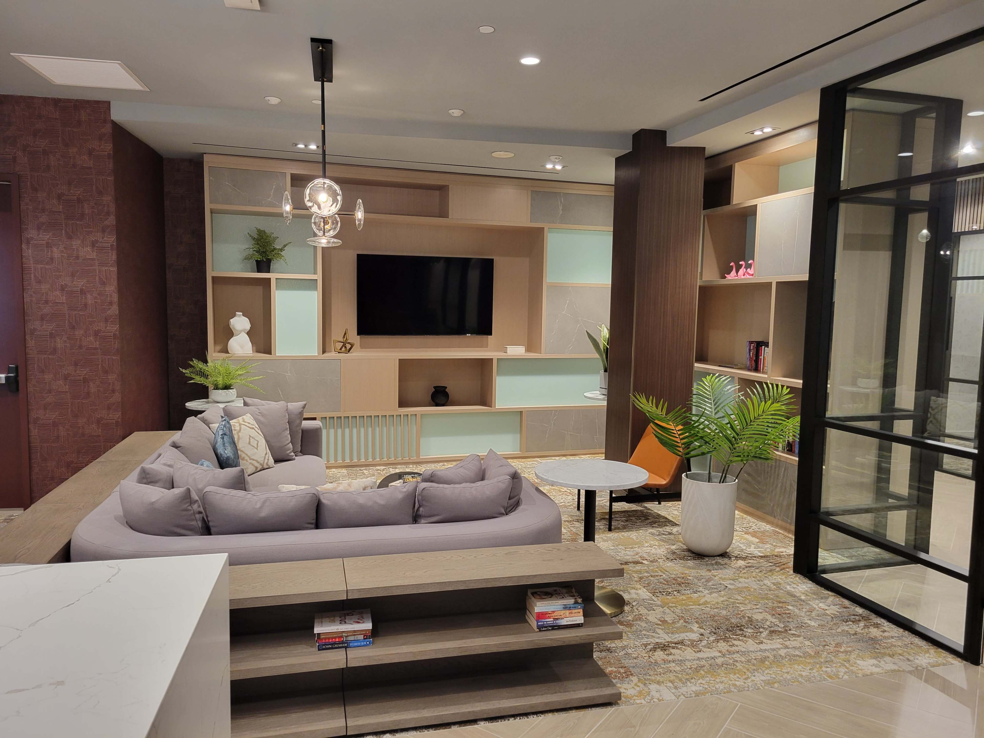 A modern lounge area with a sectional sofa, a coffee table, a large television mounted on a wall unit, and decorative plants.