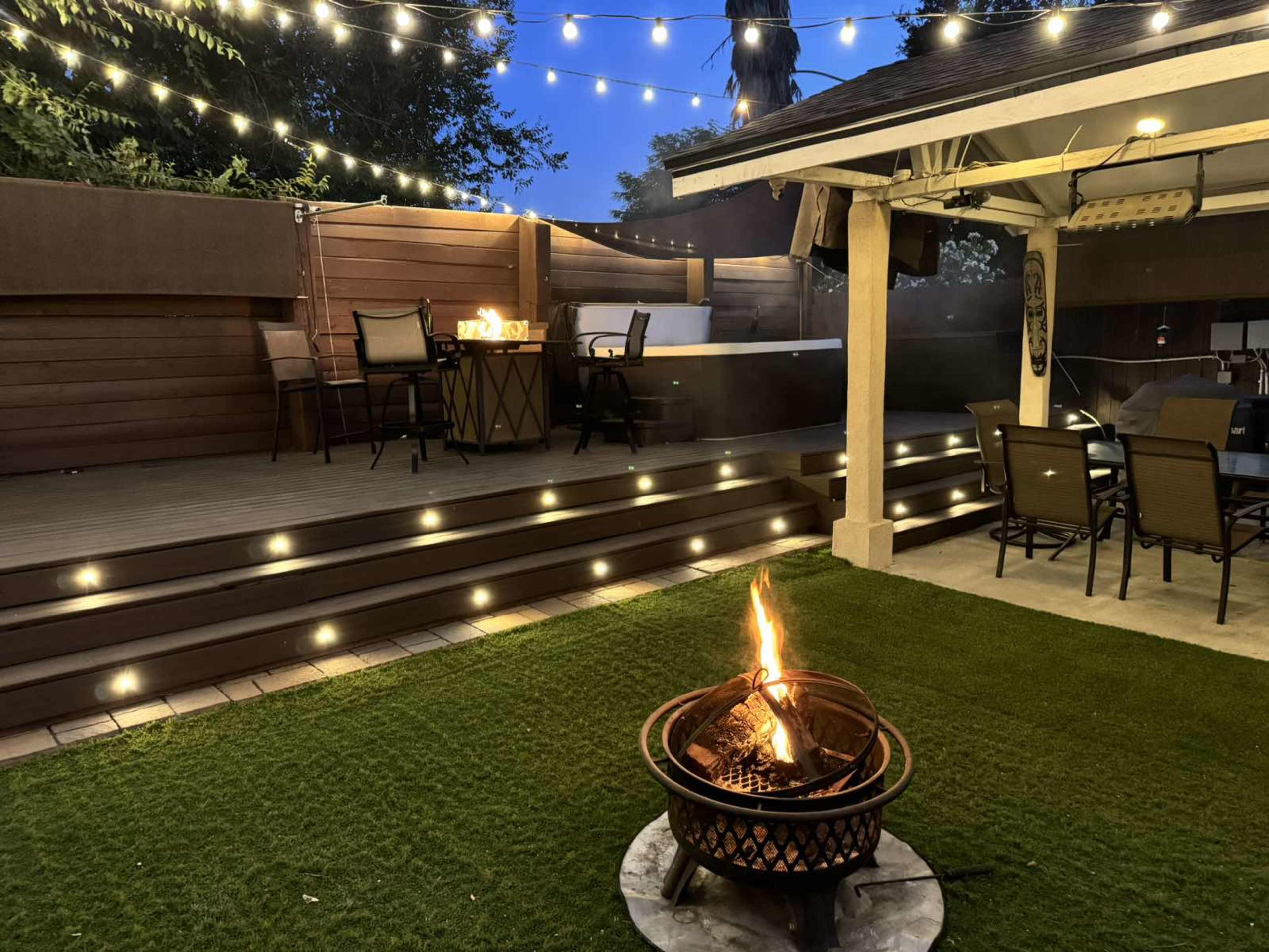 The scene features a fire pit with a flame in the foreground, surrounded by a grassy area, while string lights illuminate a wooden deck with outdoor seating in the background.