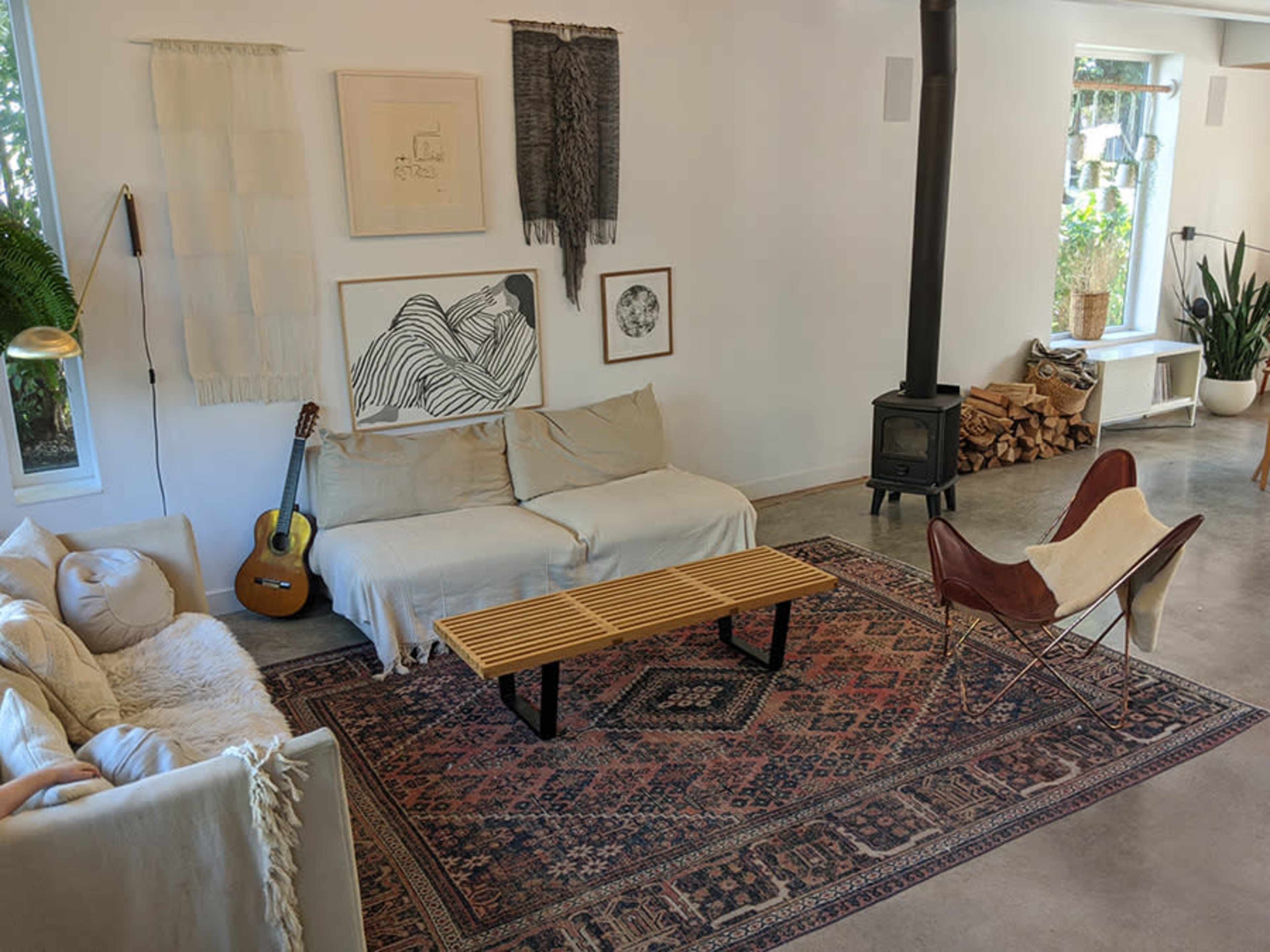 The image shows a living room with a beige sofa, a wooden coffee table, an acoustic guitar, a patterned rug, and a wood stove, surrounded by plants and wall art.