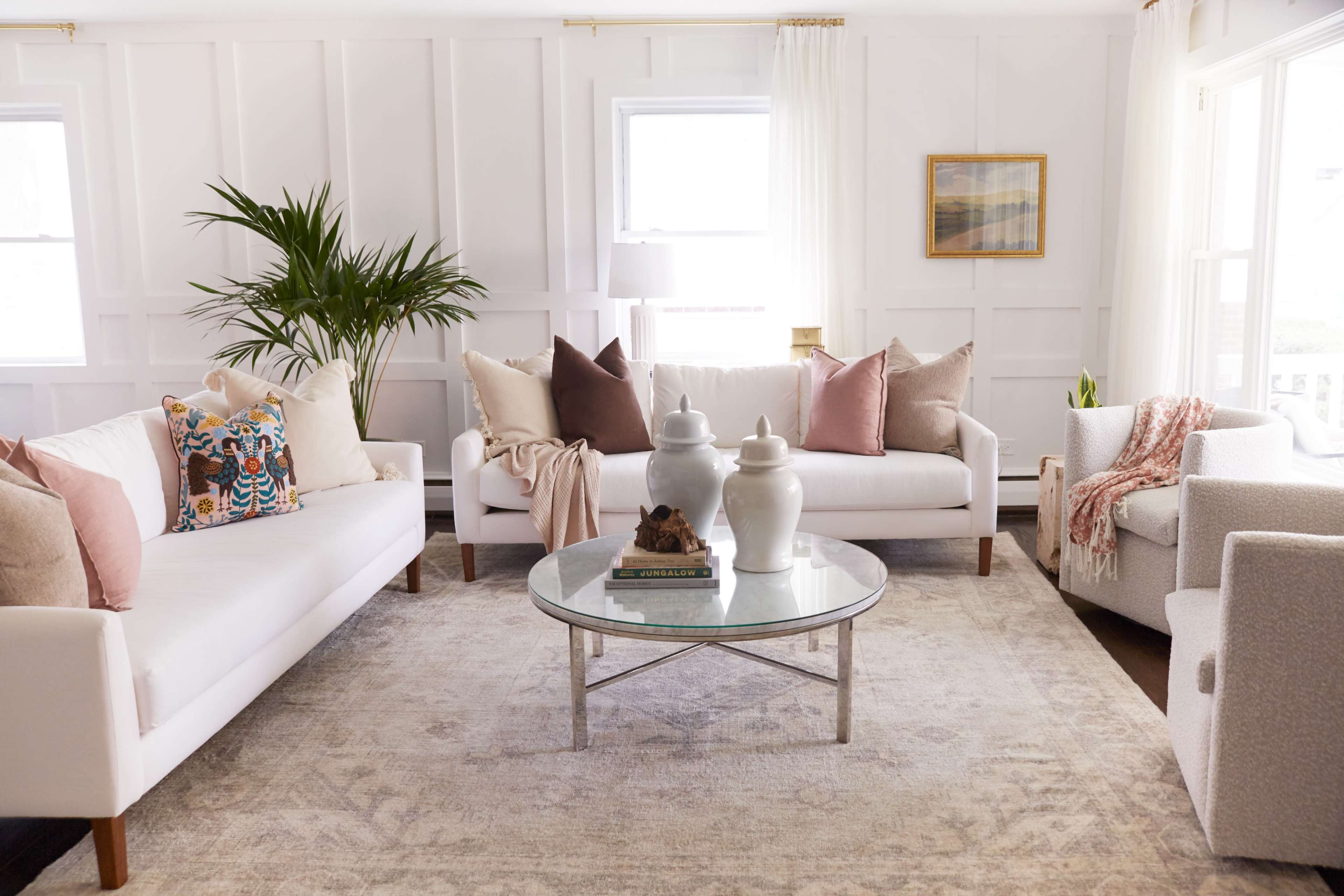 A bright, stylish living room features two white sofas with decorative pillows, a glass coffee table, and a soft area rug, surrounded by plants and natural light.