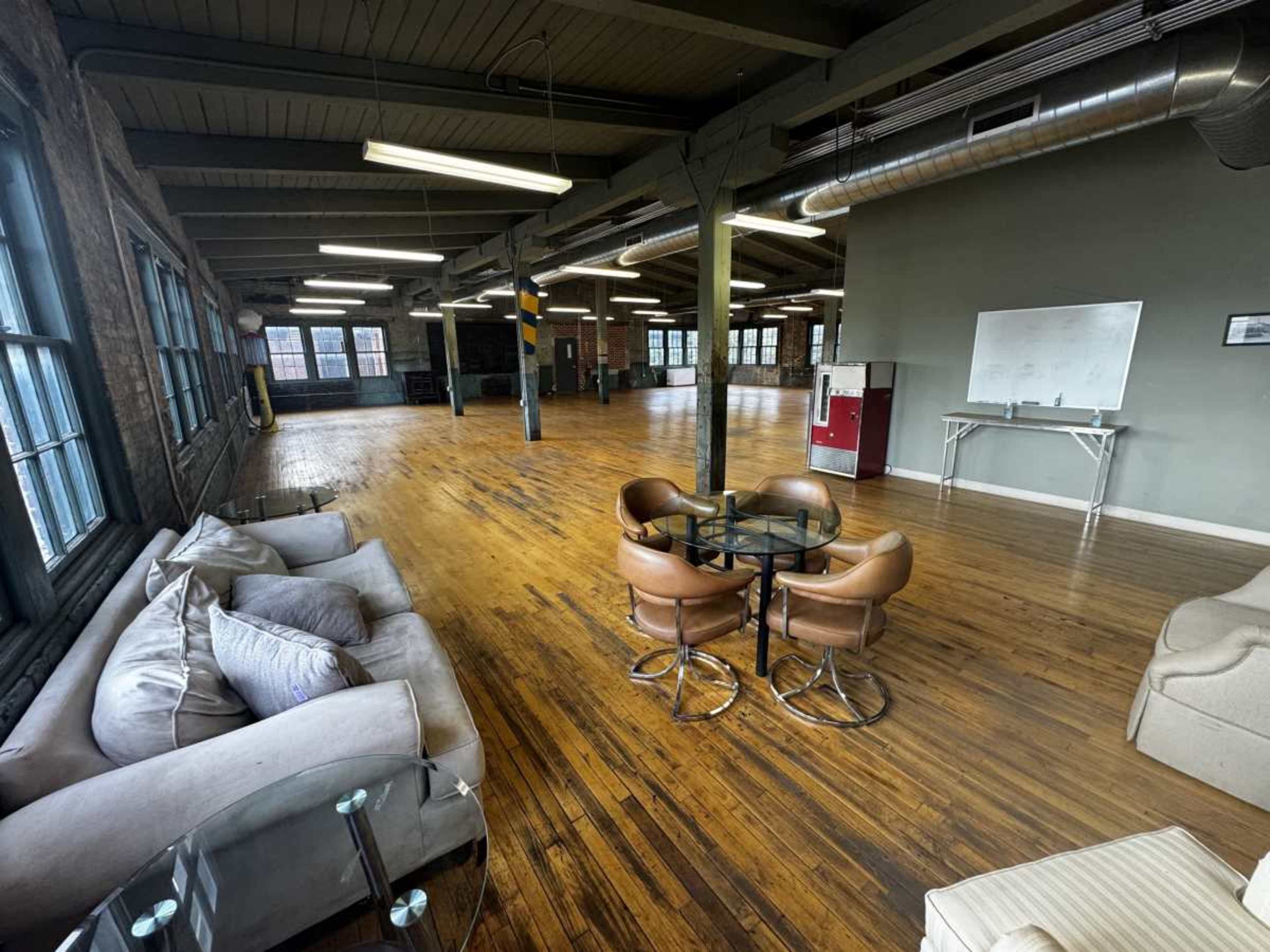 The image shows a spacious, industrial-style room with wooden flooring, large windows, and seating arrangements that include a glass table and two brown chairs.