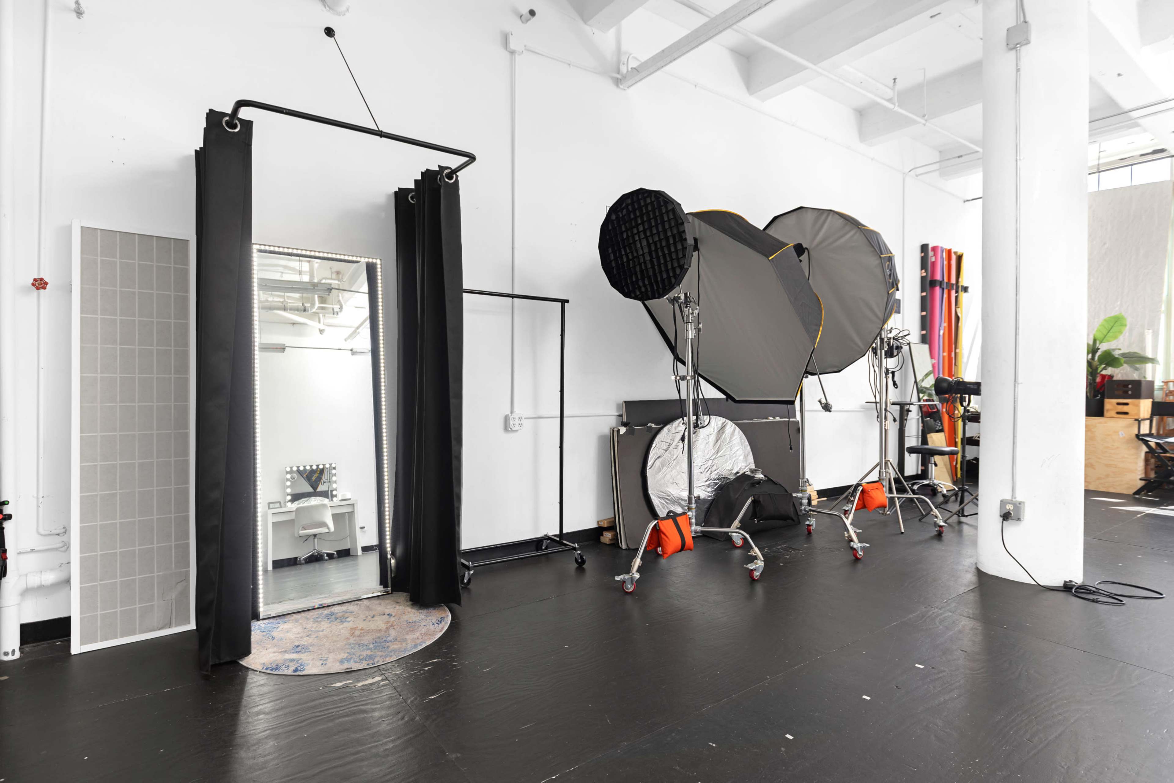 The image shows a spacious studio interior with a large mirror, photography lighting equipment, and a minimalist black floor.