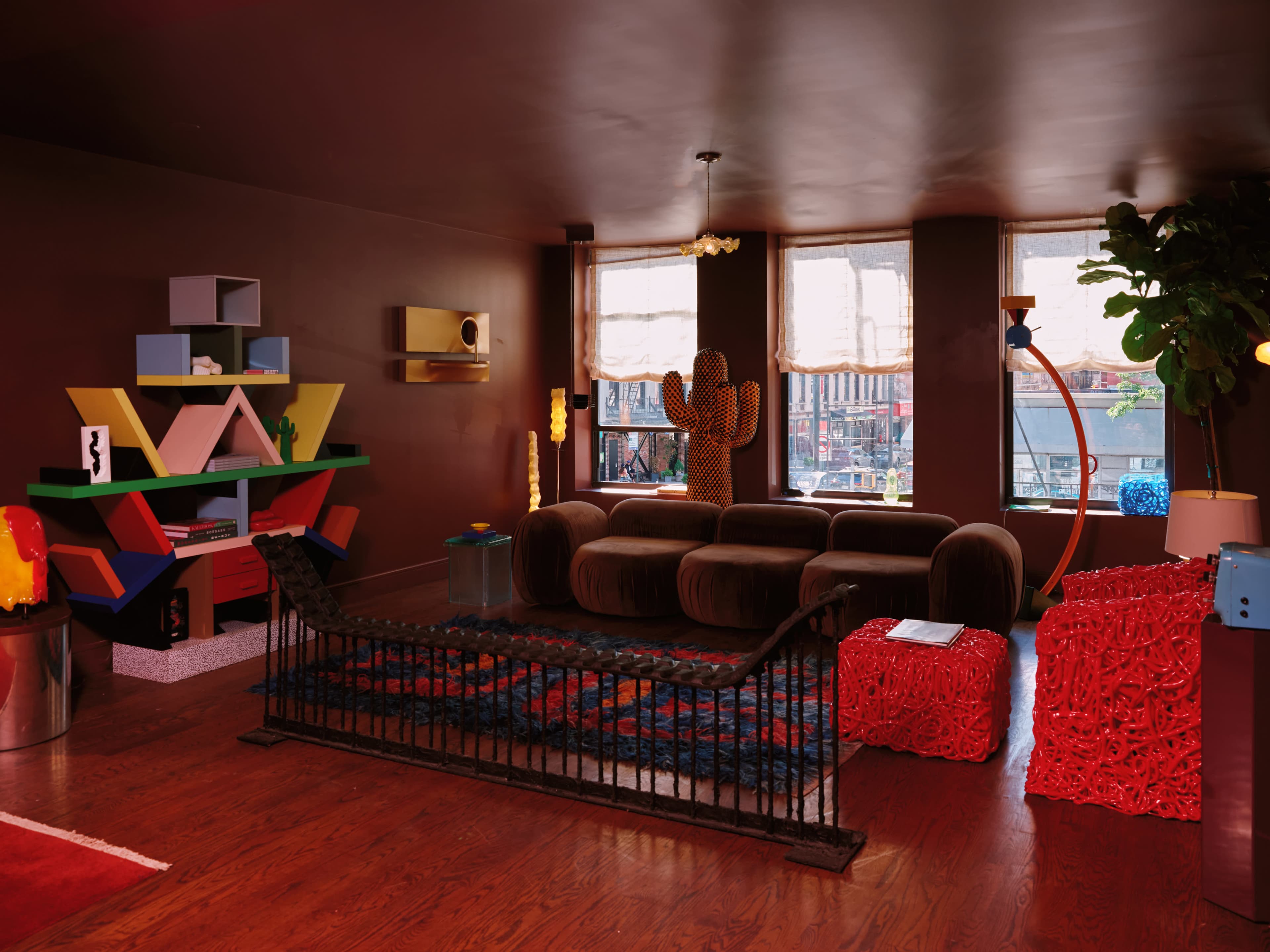 The image shows a living room with a brown sofa, colorful shelving unit, vibrant decor, and large windows allowing natural light.