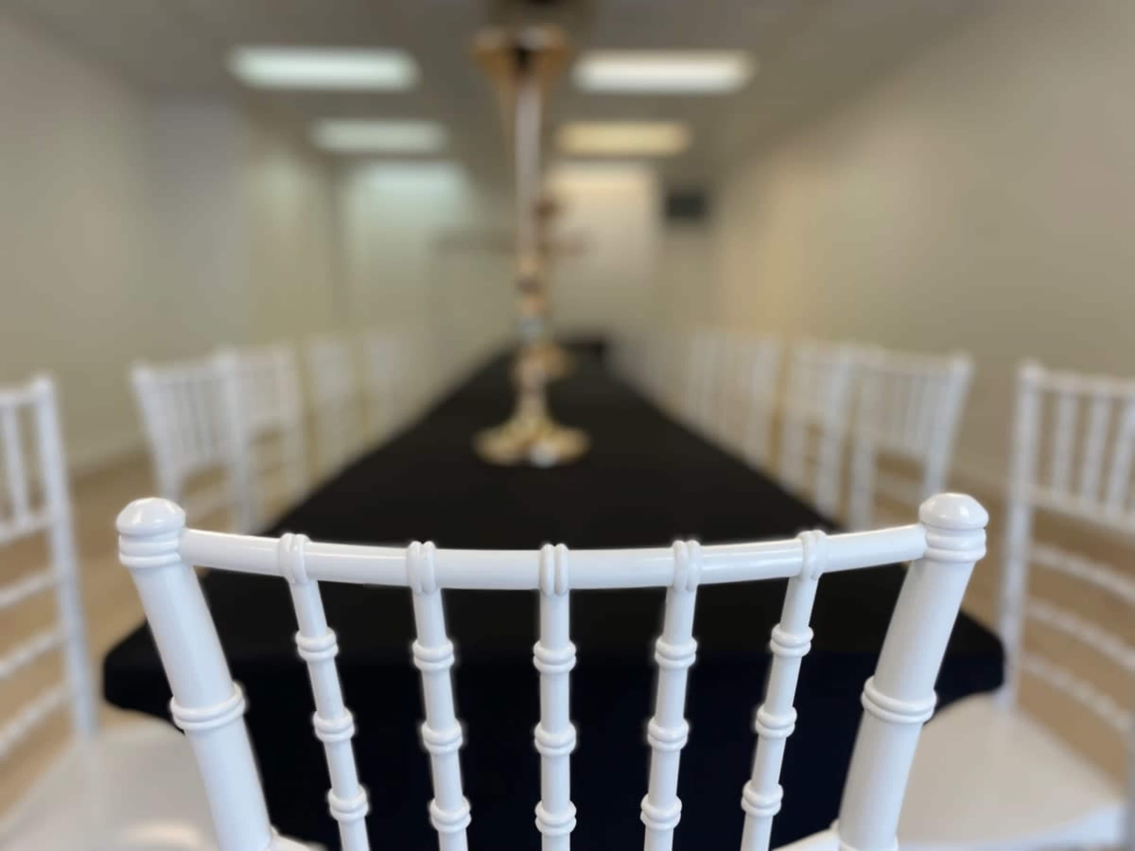 A long, black table is set with white chiavari chairs arranged around it in a well-lit room.
