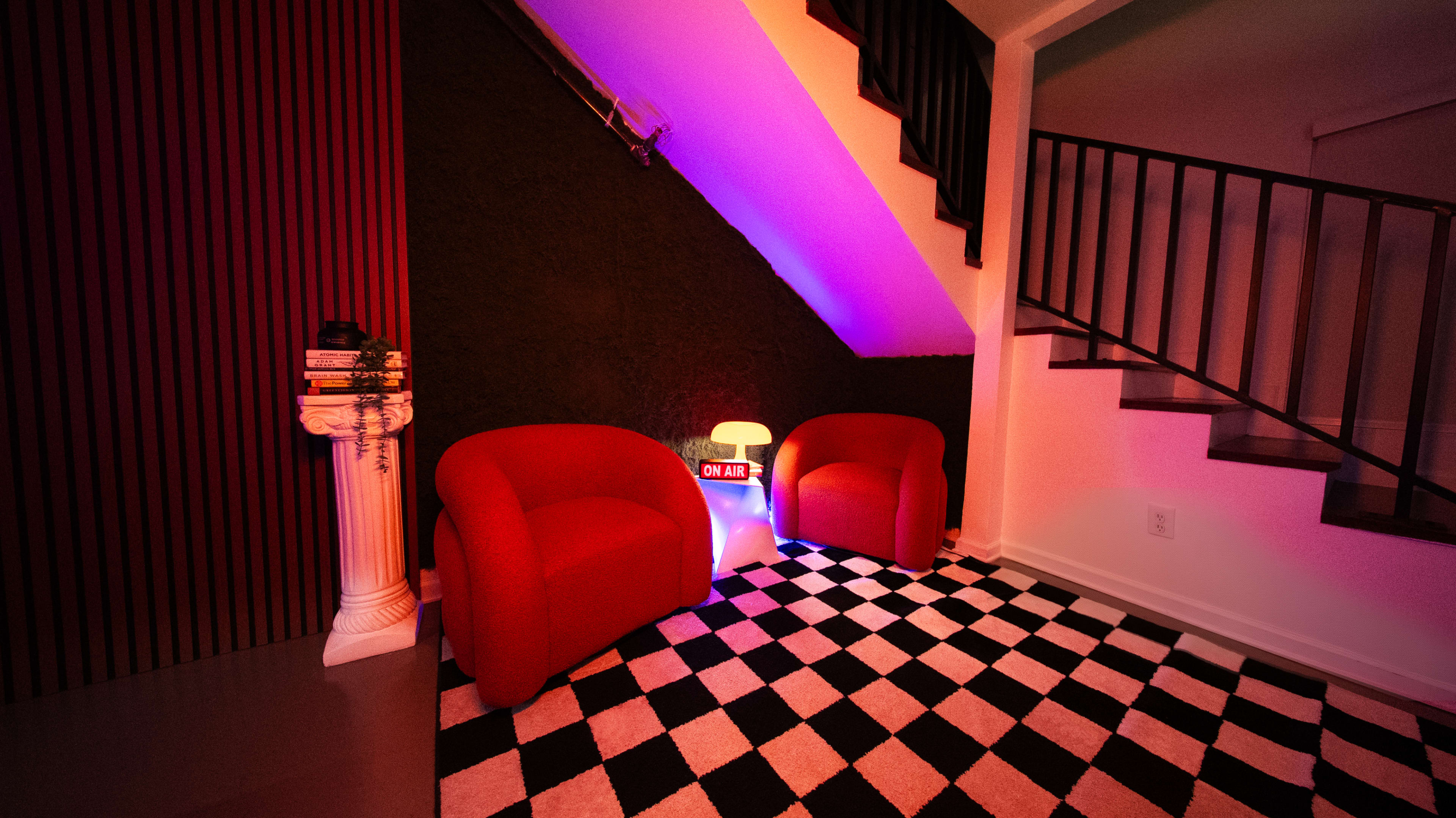 The scene features two red chairs positioned on a black-and-white checkered rug, with a decorative column and a lamp displaying "ON AIR" in a well-lit corner beneath a staircase.