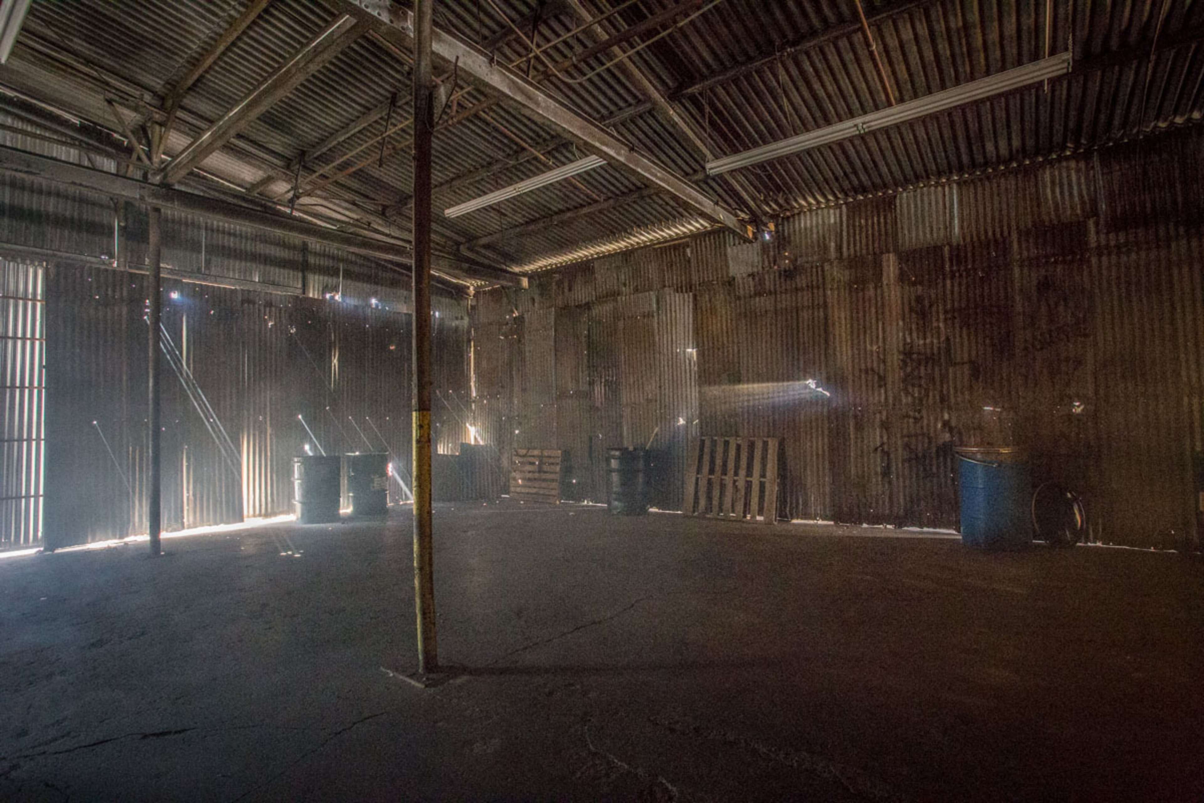 The image shows a dimly lit industrial space with corrugated metal walls, featuring scattered barrels and wooden pallets.