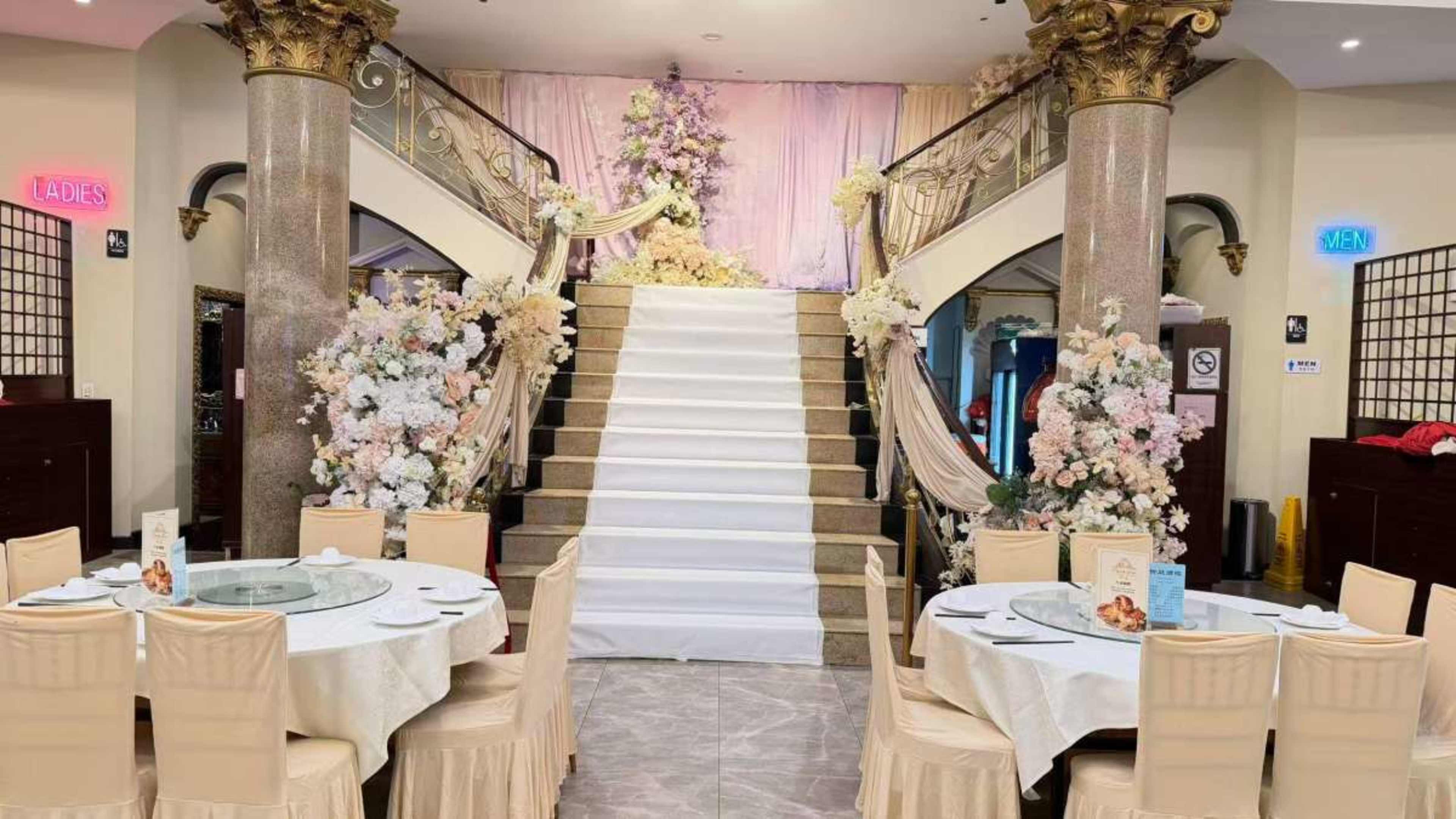 The image shows a large, elegantly decorated dining area featuring a staircase adorned with flowers and fabric draping, surrounded by round tables set with white tablecloths.
