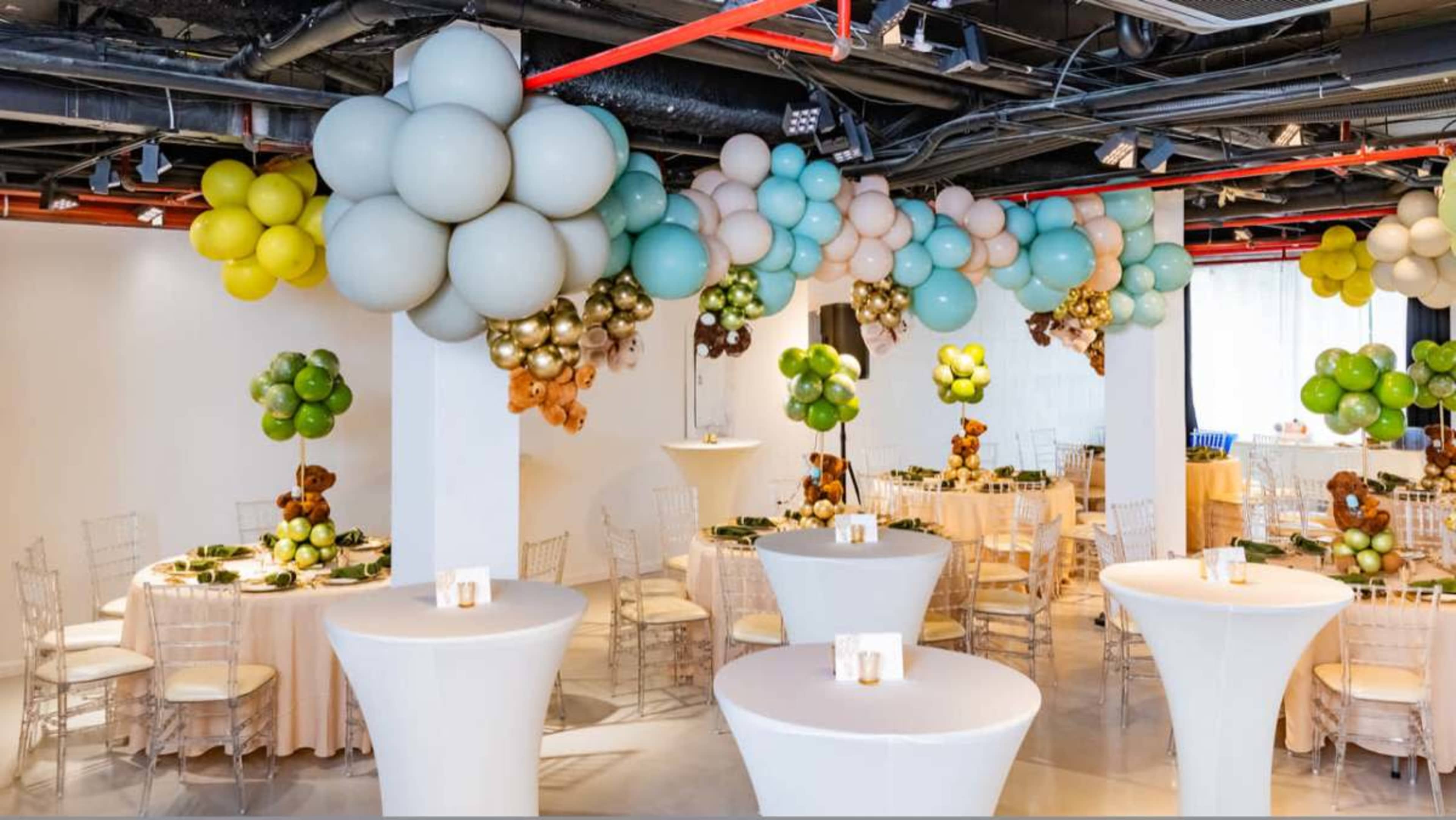 A decorated event space features elegant tables with balloon arrangements in green and blue tones overhead.