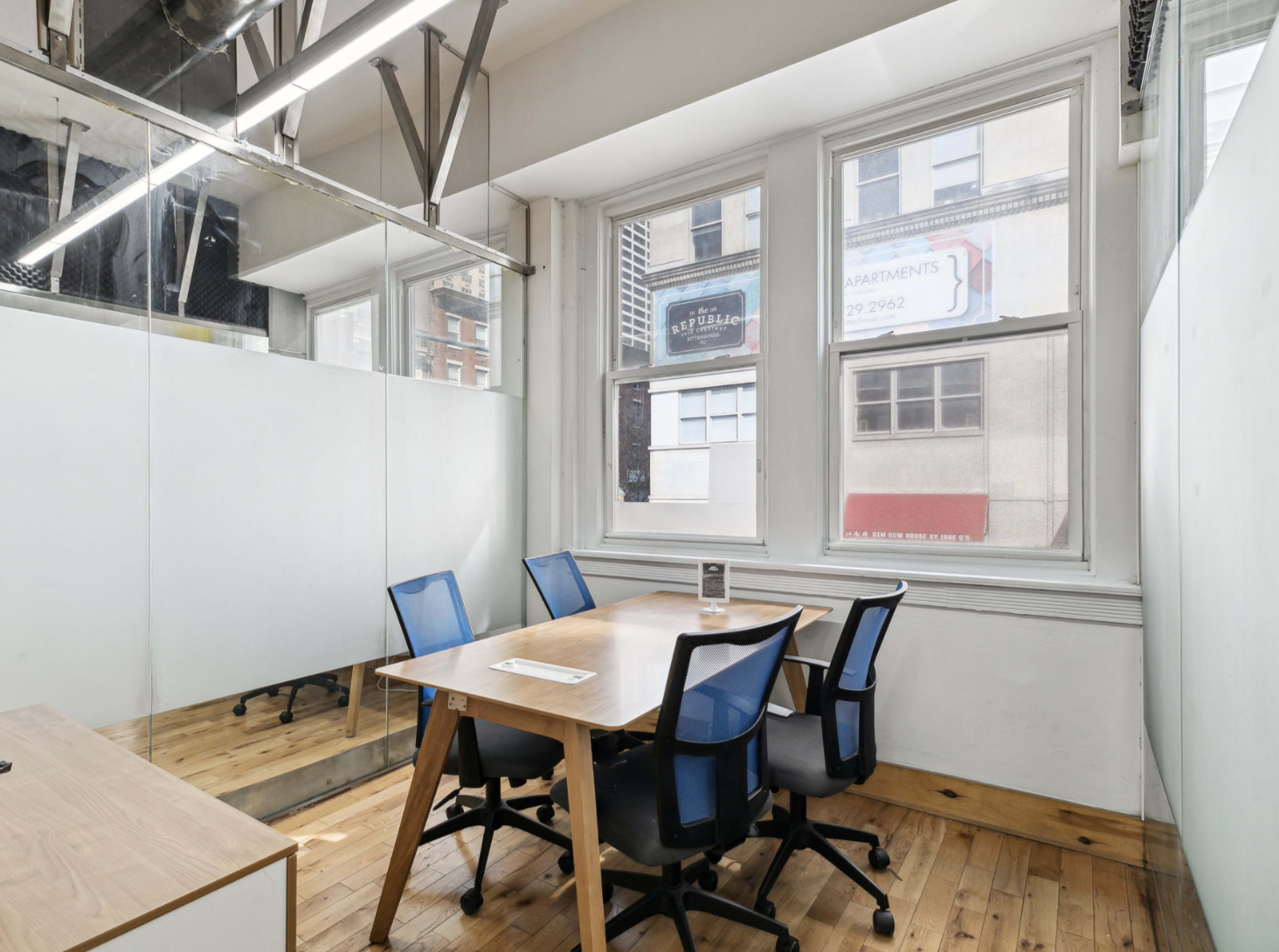 The image shows a small meeting room featuring a wooden table with four blue chairs, large windows displaying an urban view, and a glass partition on one side.