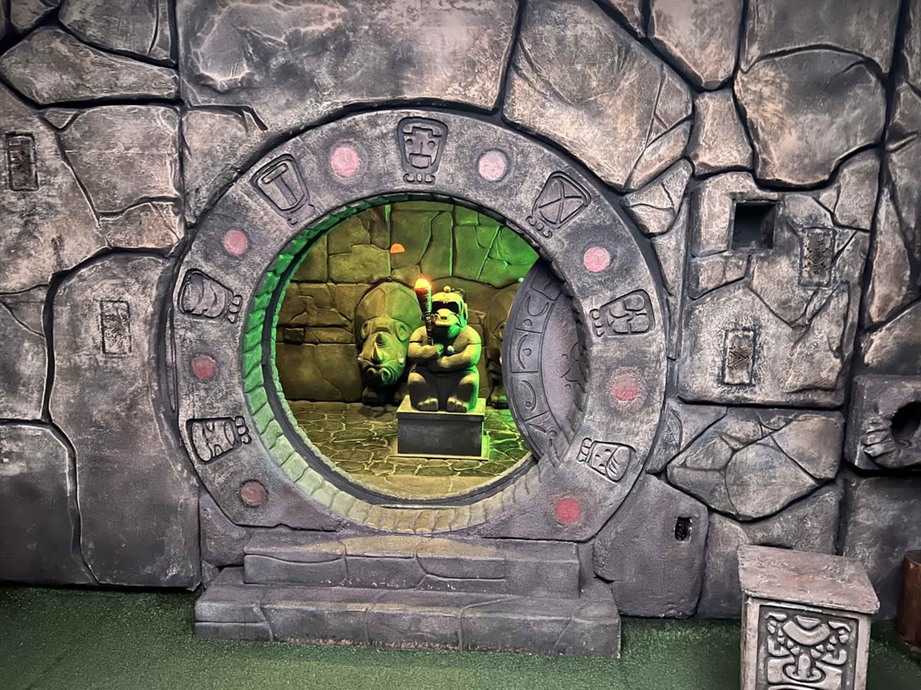 The image shows a stone archway with an intricately carved circular frame, leading to a room featuring a statue of a figure and a bear against a textured rock wall.