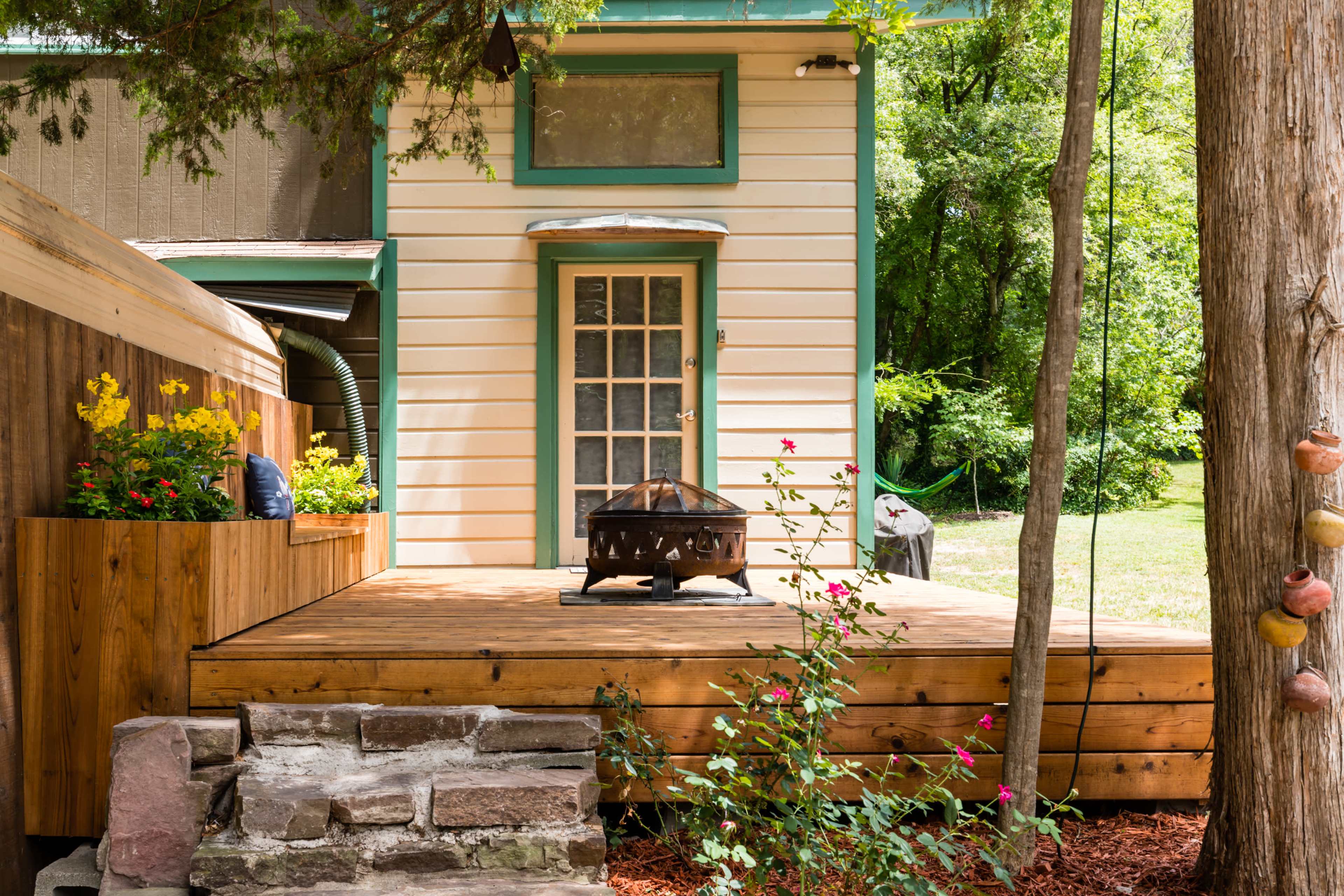 A wooden deck with a fire pit is situated in front of a yellow house with green trim, surrounded by trees and garden plants.