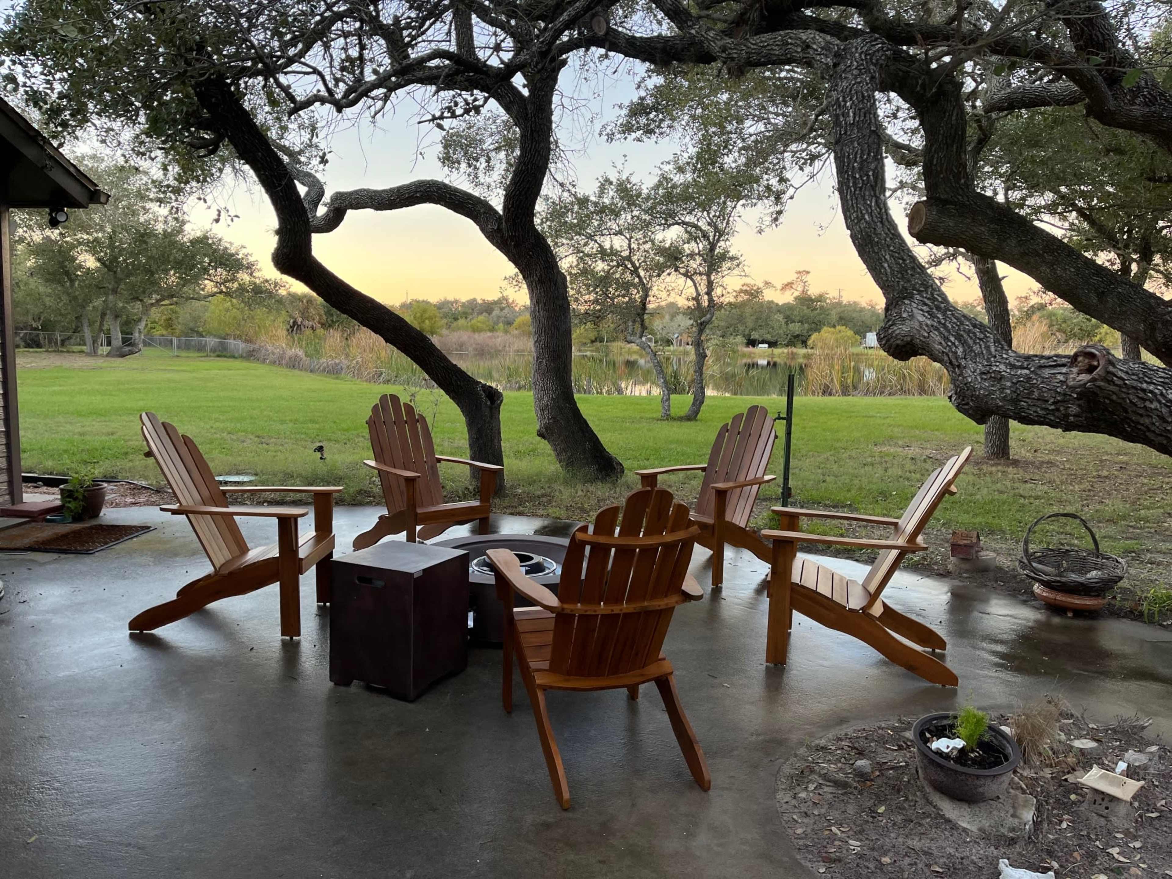 A patio area features five wooden Adirondack chairs arranged around a small fire pit, under large trees, with a view of a waterway in the background.