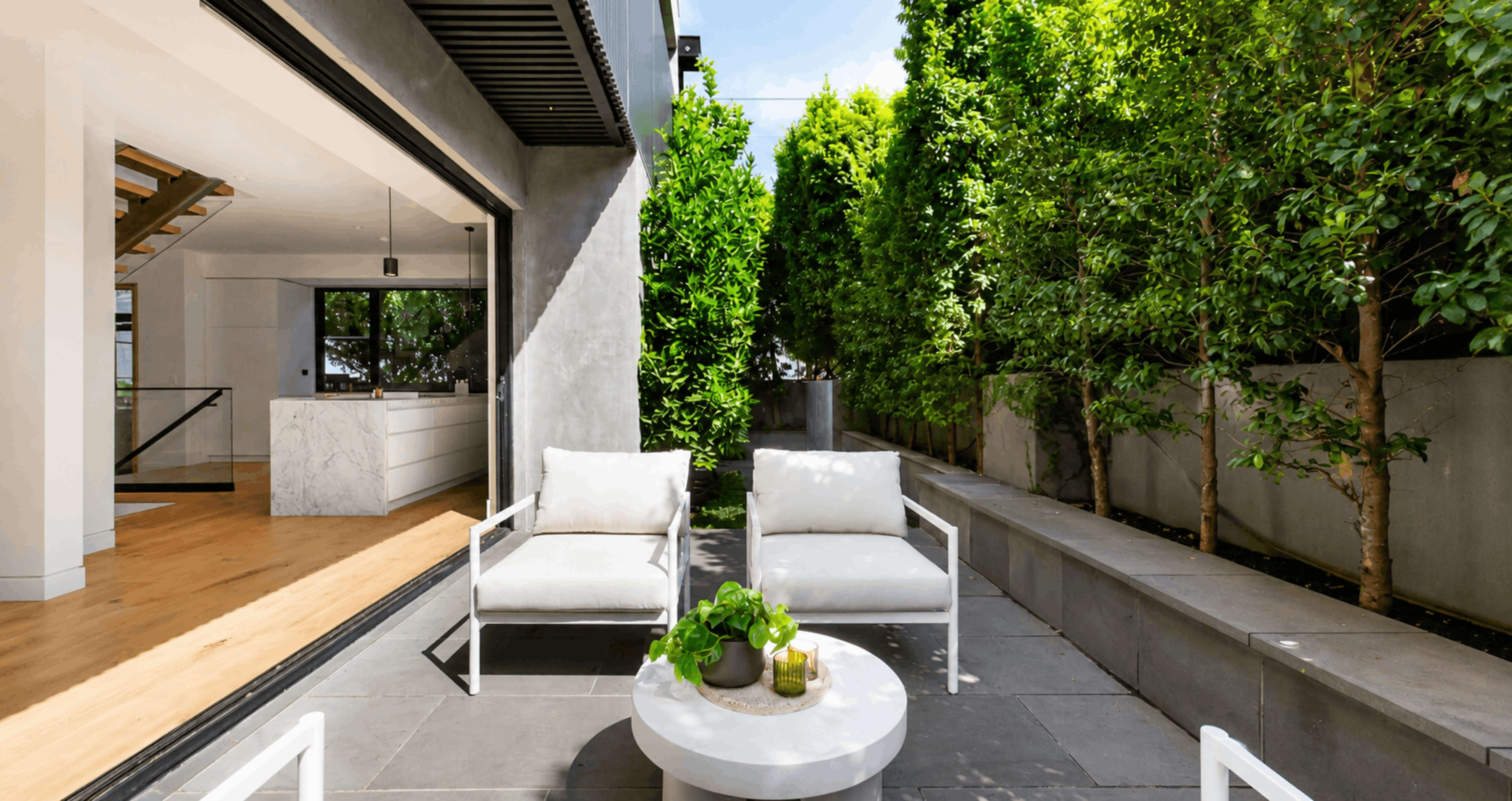 A modern outdoor seating area features two white chairs and a round table nestled between tall green hedges and the interior of a contemporary home.