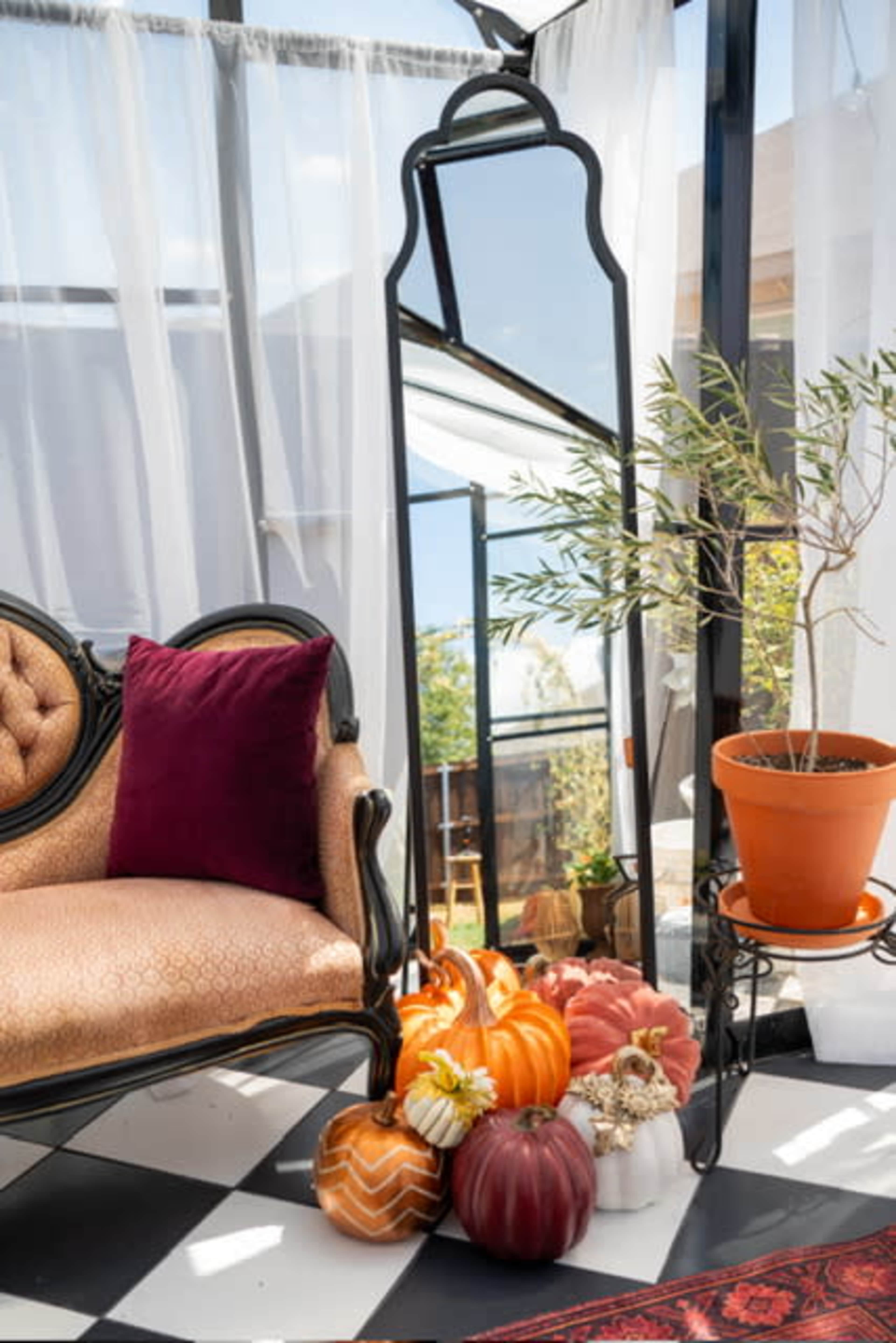 A vintage sofa with a burgundy pillow is positioned next to a large mirror and decorated with various pumpkins in a bright, sunlit space.